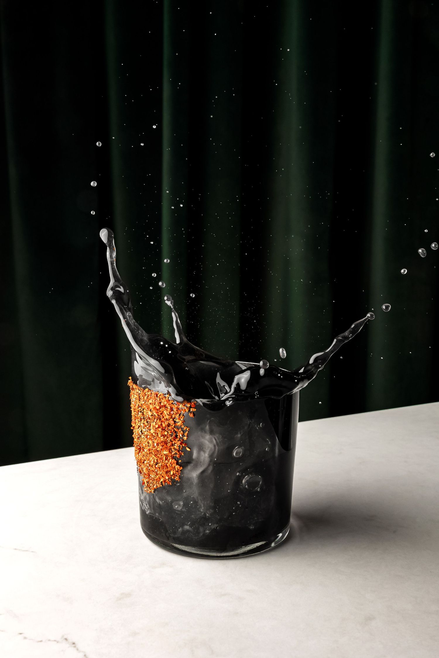 A dramatic splash of dark liquid in a glass with an orange-rimmed edge against a black background.