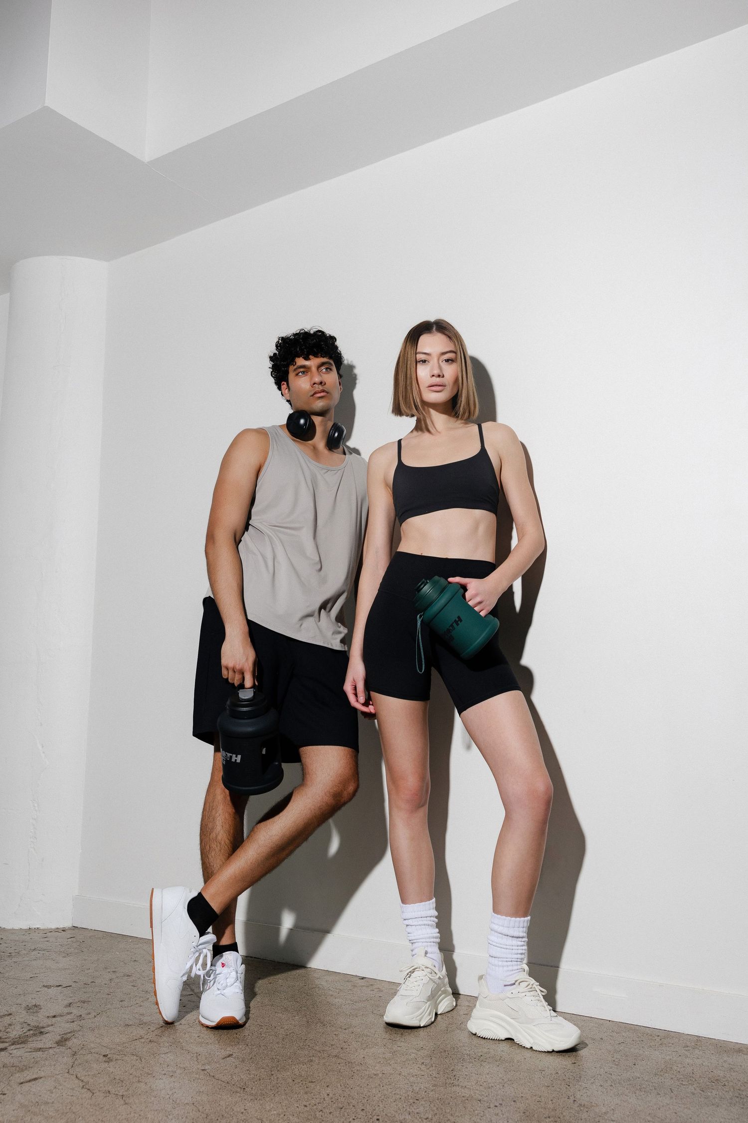 Two people in workout attire sitting against white wall with fitness accessories.