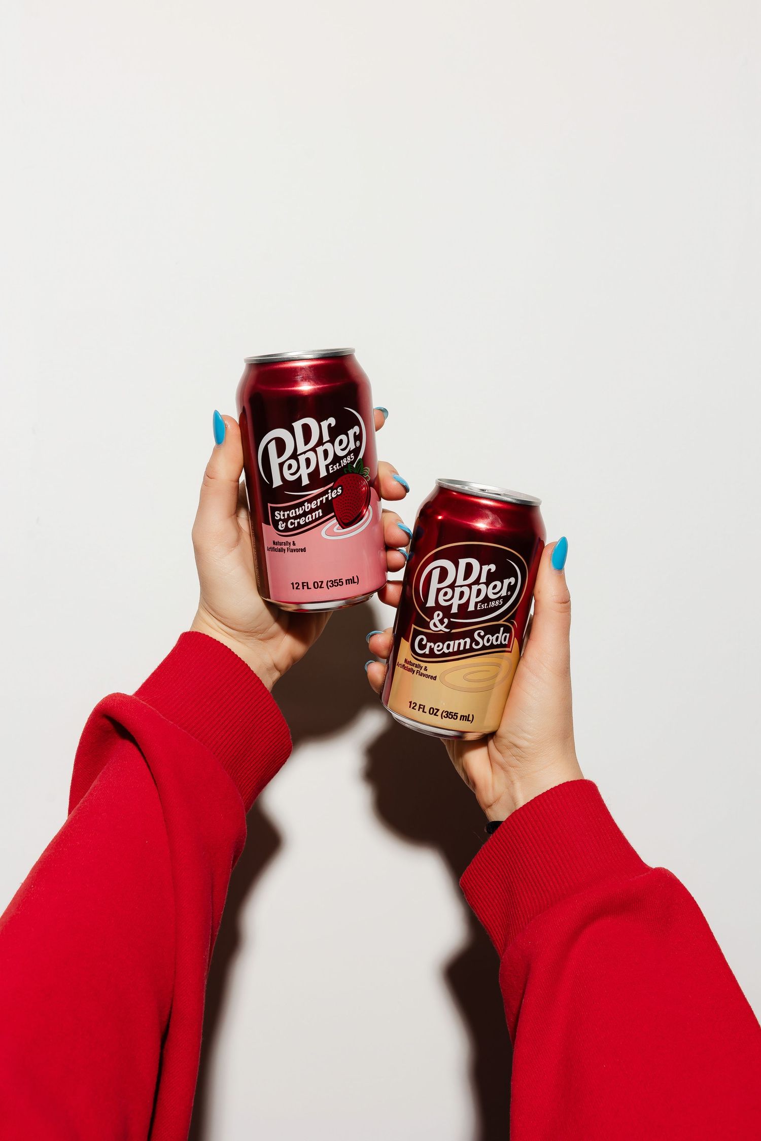Hands holding Dr Pepper cans against white background wearing red sleeves.