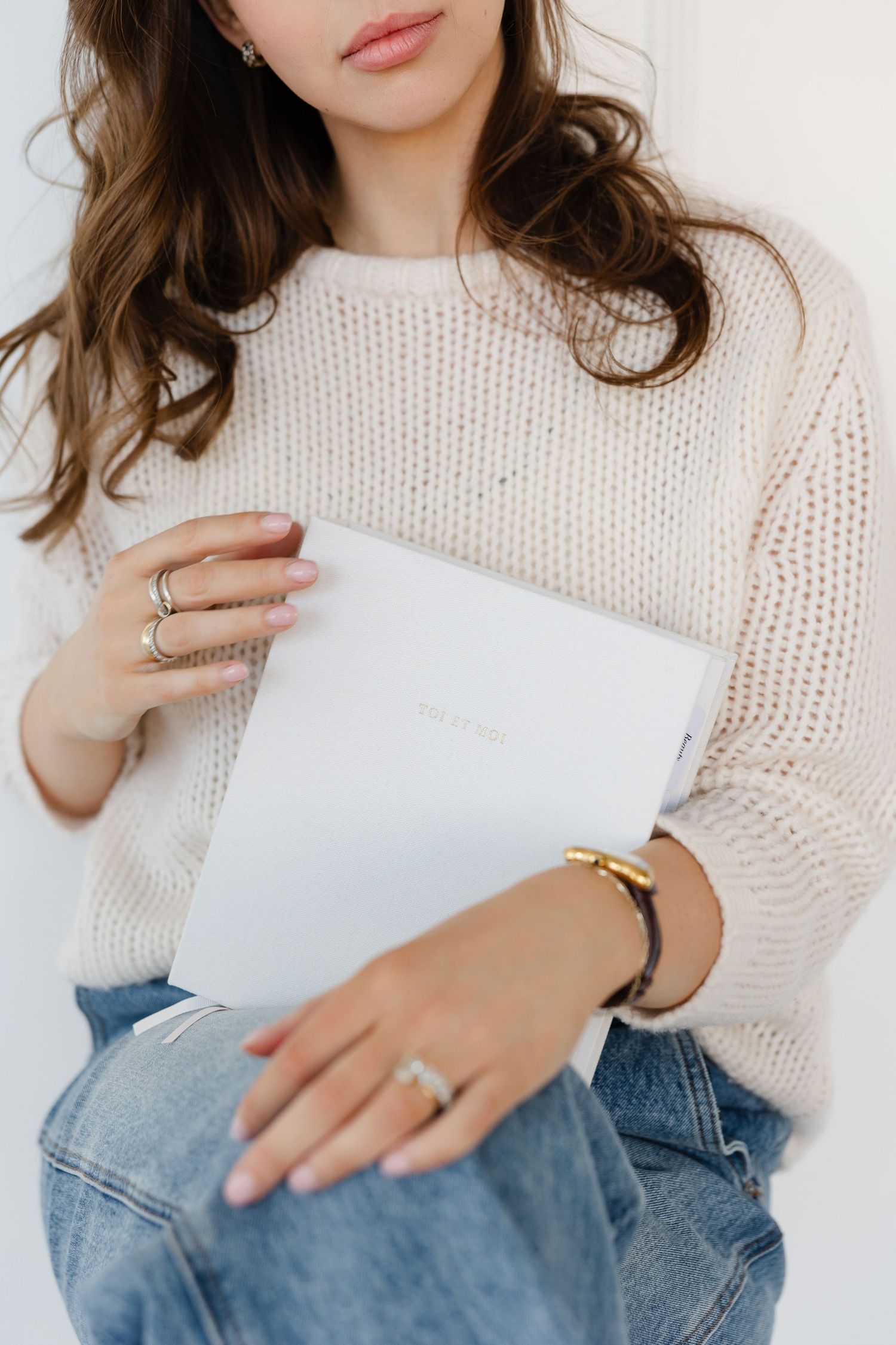 Close-up of hands with rings holding white book, wearing cream knit sweater.