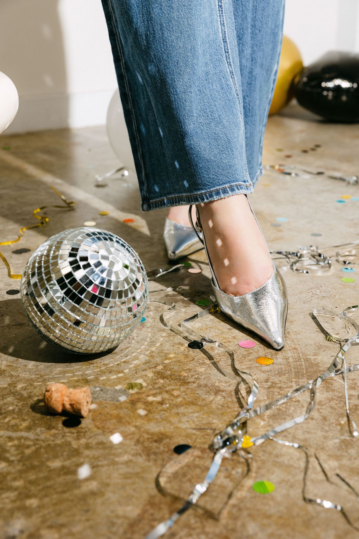 Silver metallic pointed toe heels next to disco ball with scattered confetti on ground.