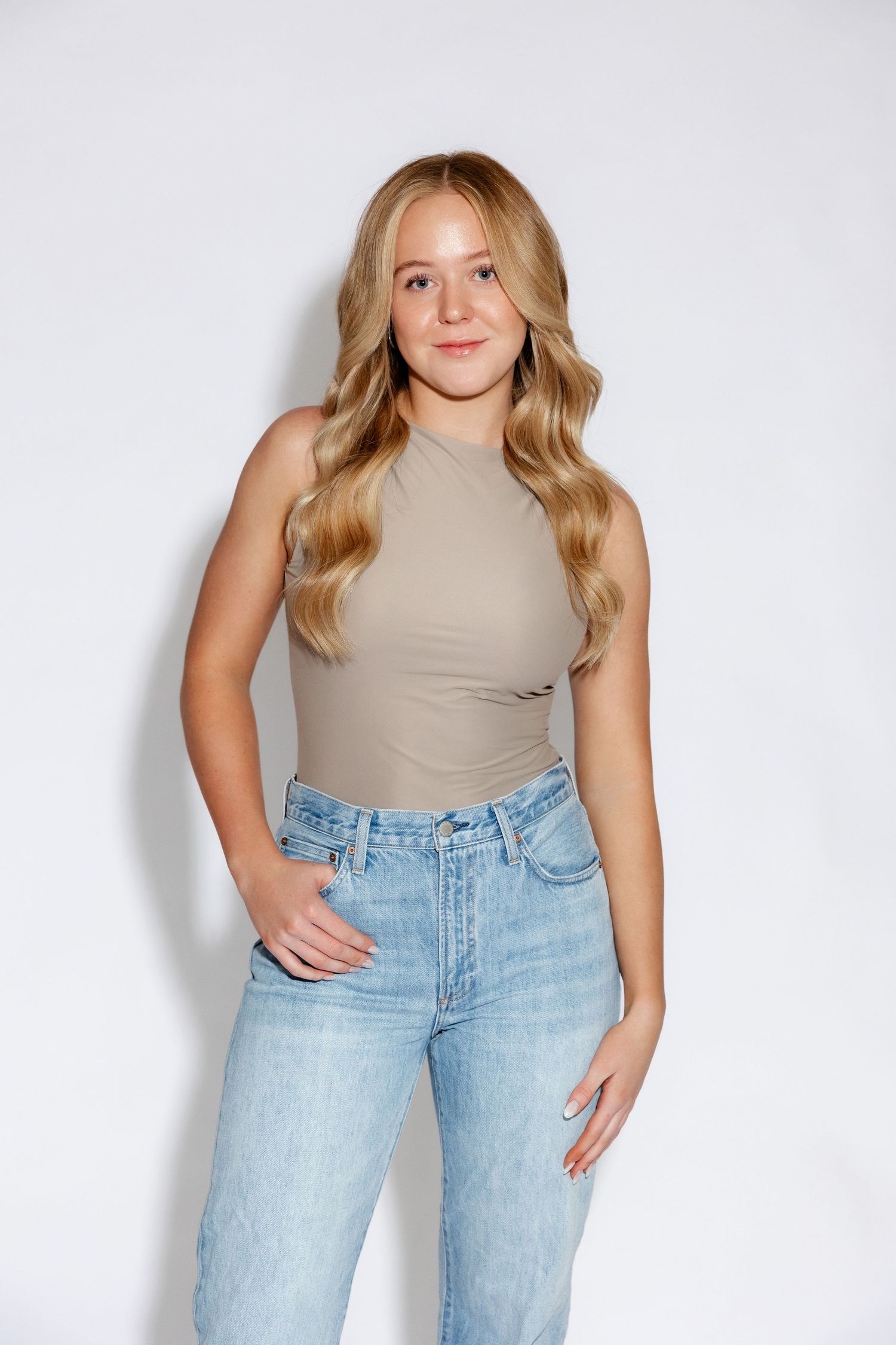 Woman in sleeveless beige top and light jeans poses against white studio background.