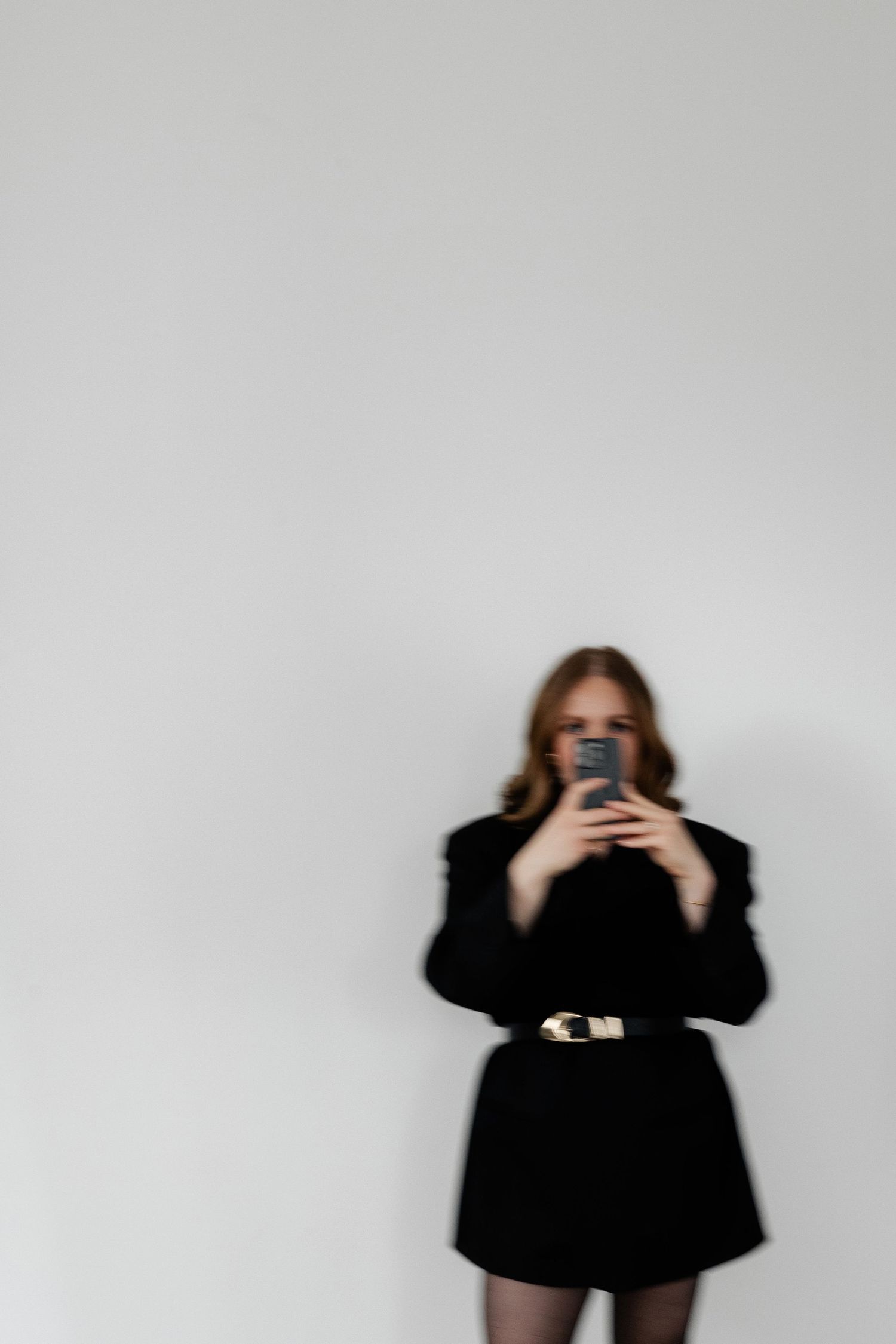 Figure in black outfit poses with phone against white wall in dramatic fashion photoshoot sequence.