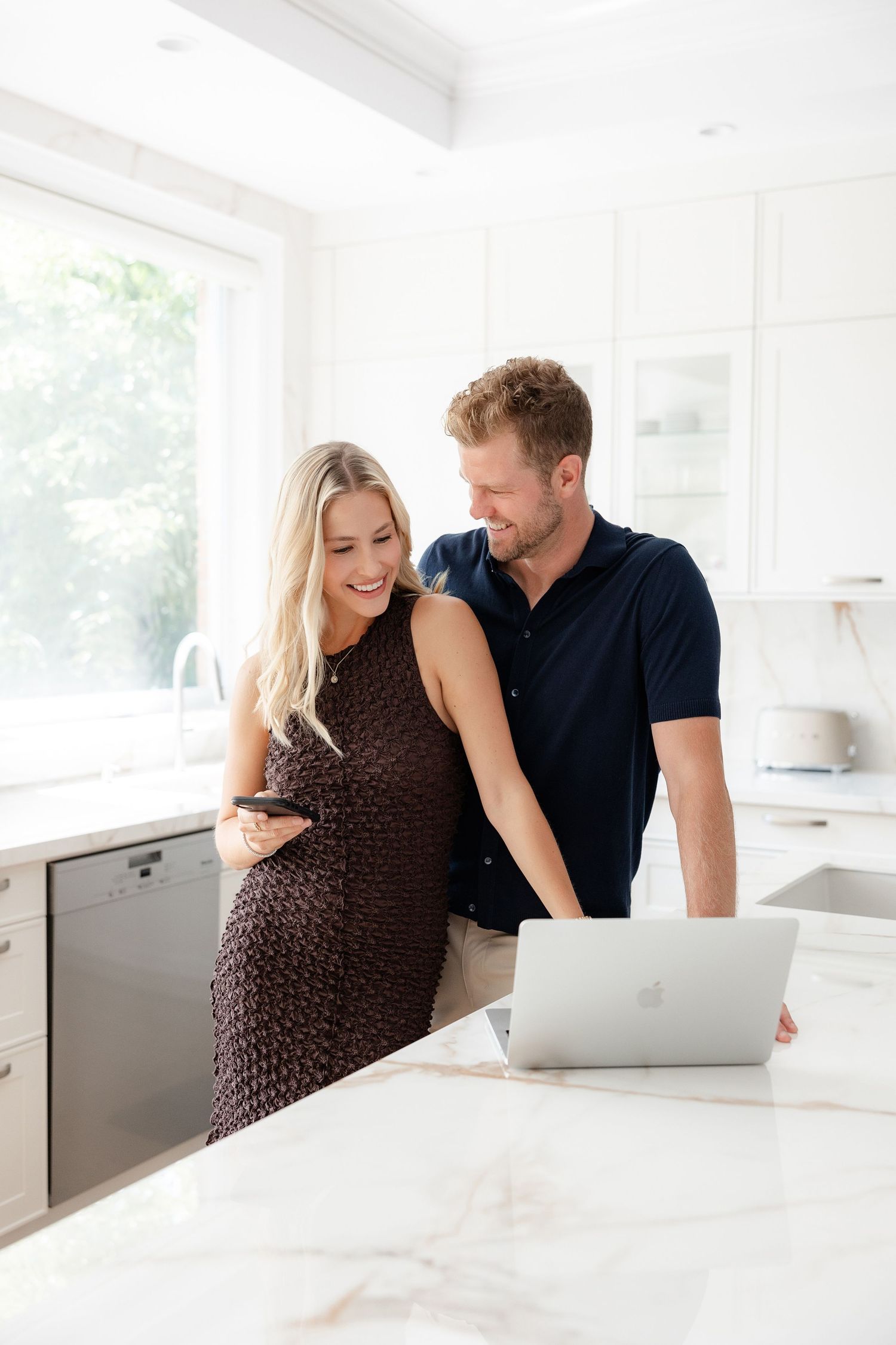 A couple reviews information together on a laptop at a white marble kitchen counter.