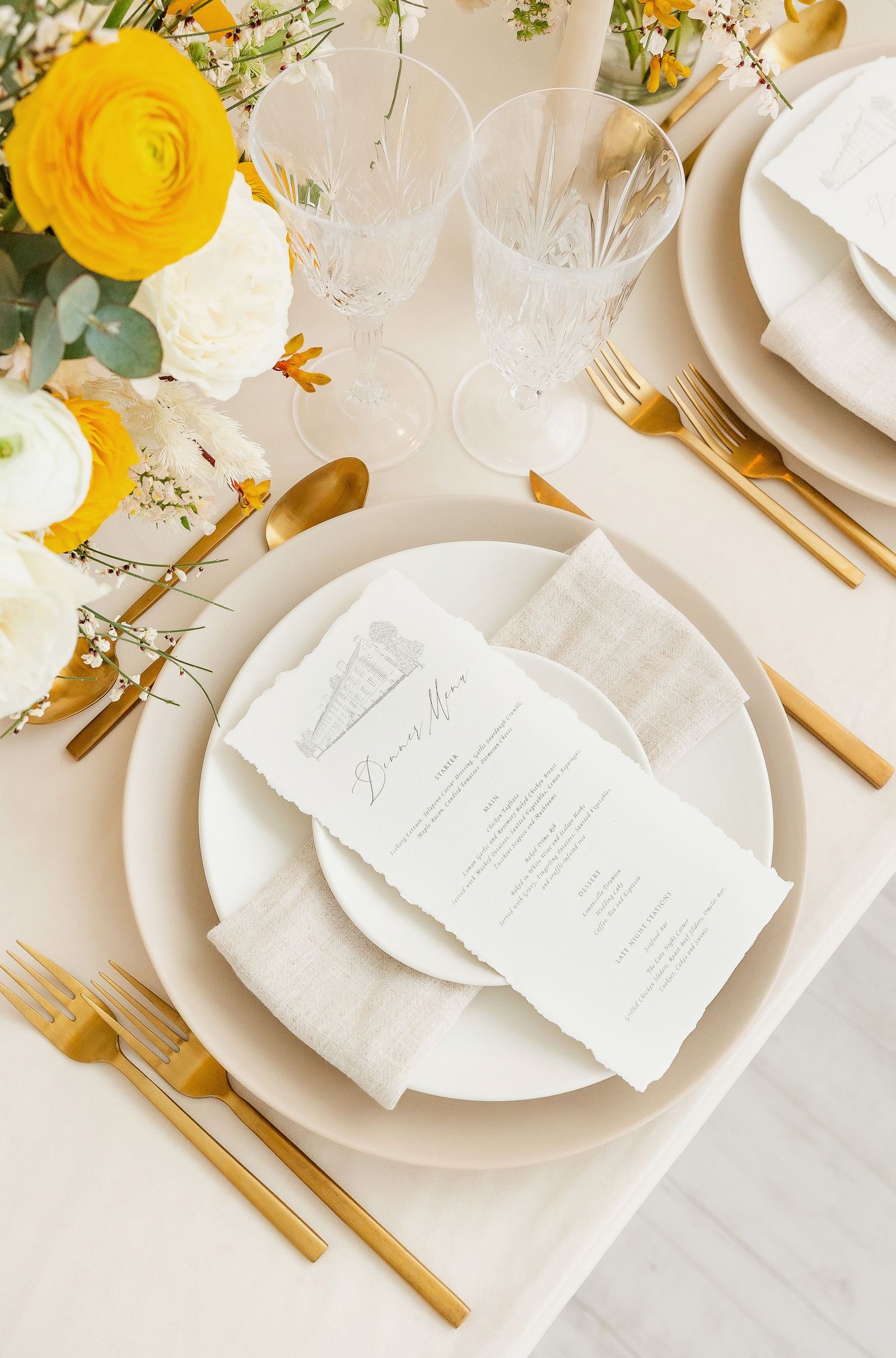 Formal dining place setting with white dishes, gold utensils, and yellow ranunculus flowers.
