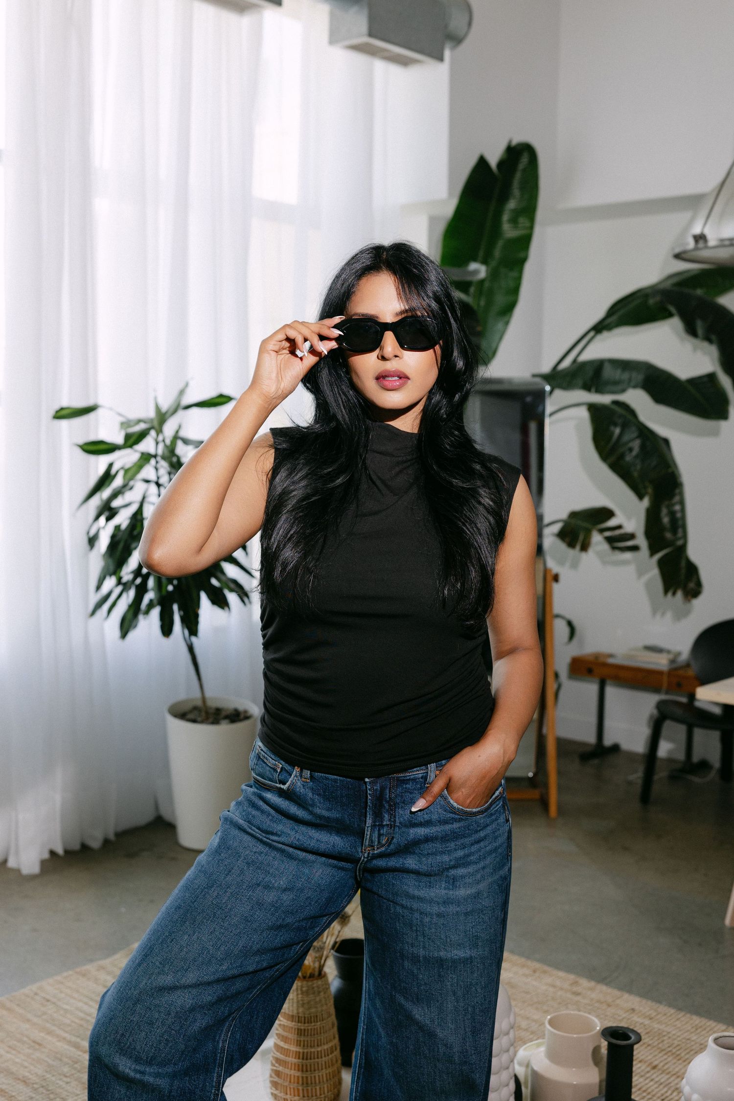 Fashion editorial in bright studio with plants showing confident poses in black top and denim jeans.