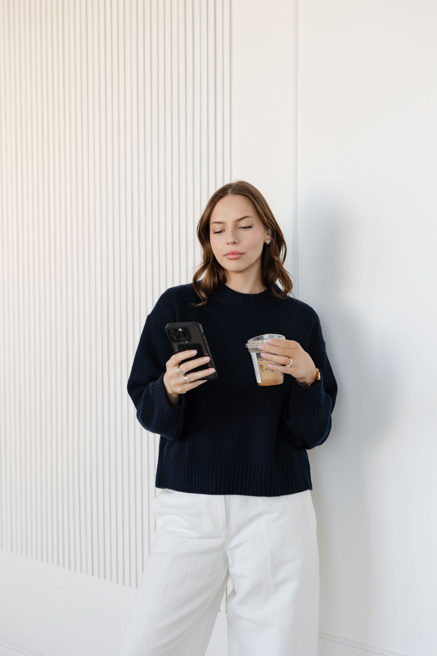 Person in black sweater using mobile phone while leaning against white wall.