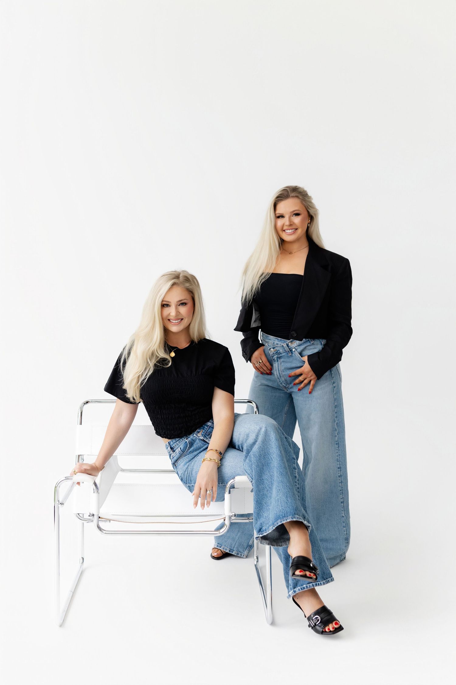 Models wearing black tops and jeans pose casually on a white bench against a bright white background.