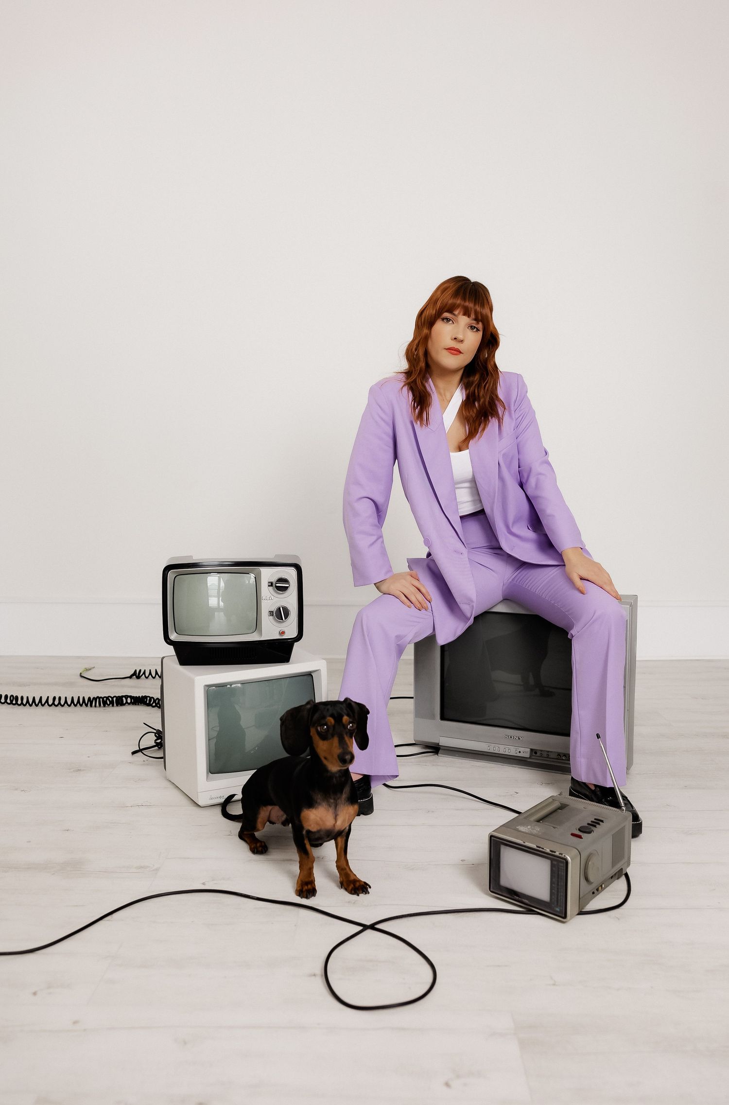 Retro-inspired photoshoot with vintage TVs, small dog, and purple pantsuit against white background.