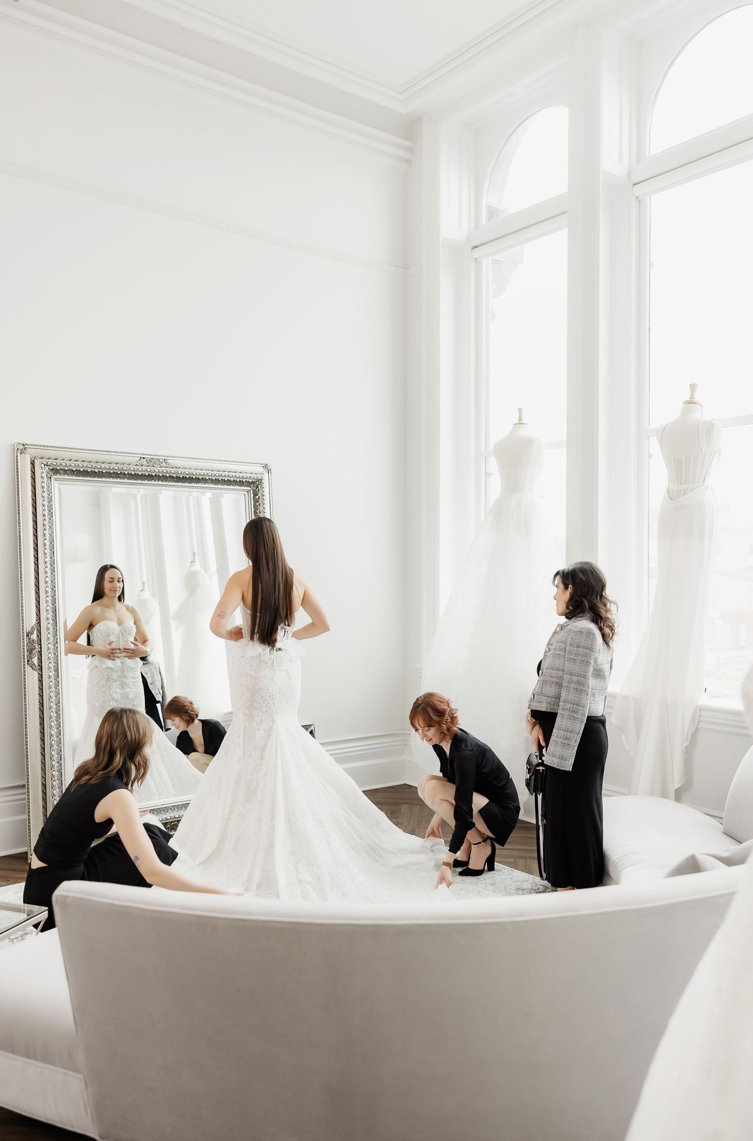 Wedding dress fitting session in an elegant white room with curved sofa and full-length mirror.