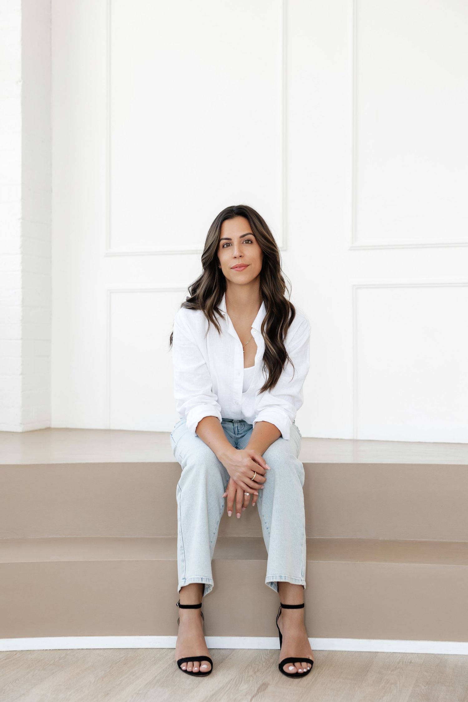 Professional woman in white shirt and light jeans sits casually on steps against white wall.