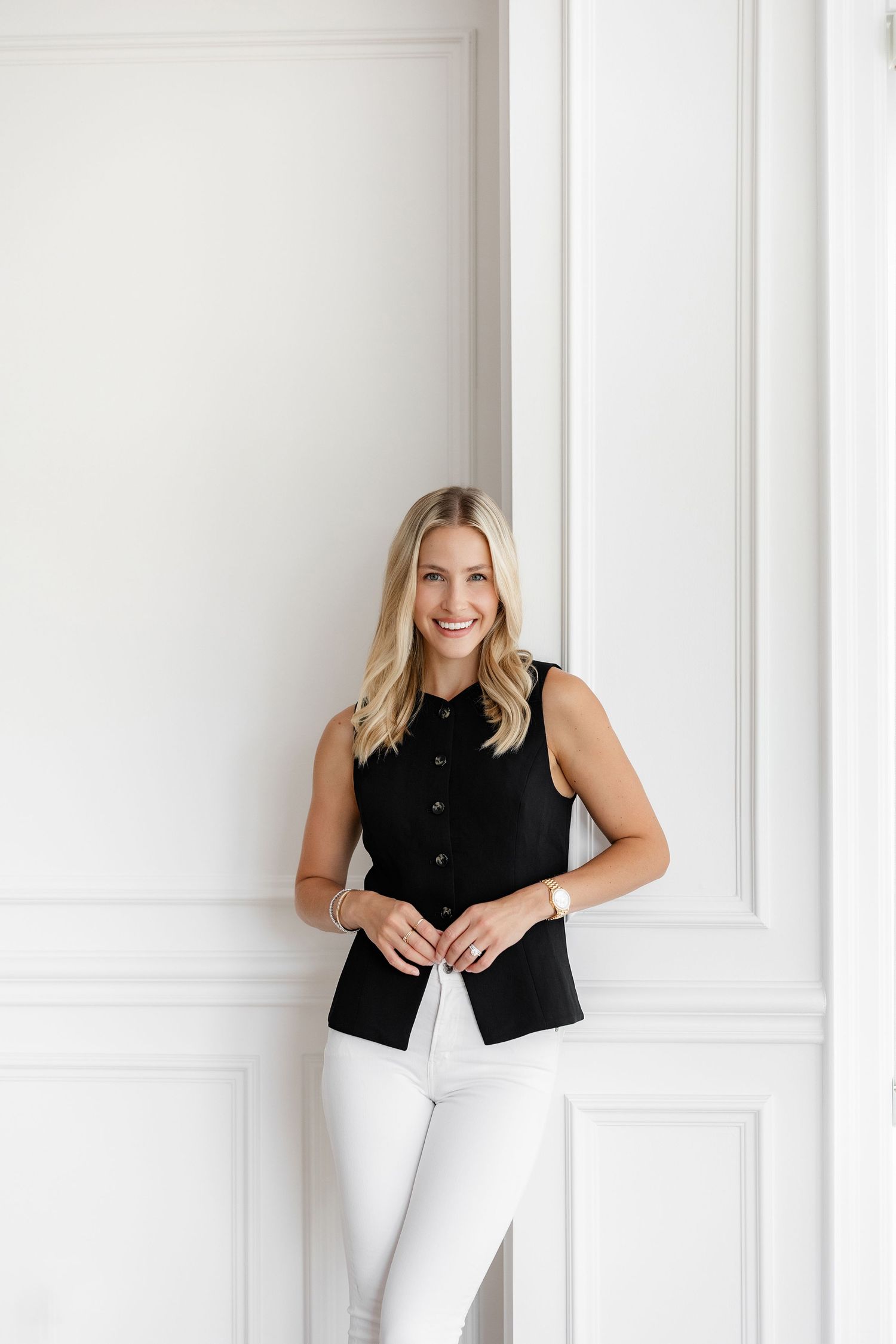 Professional portrait of person in black button-up vest and white pants against white wall.