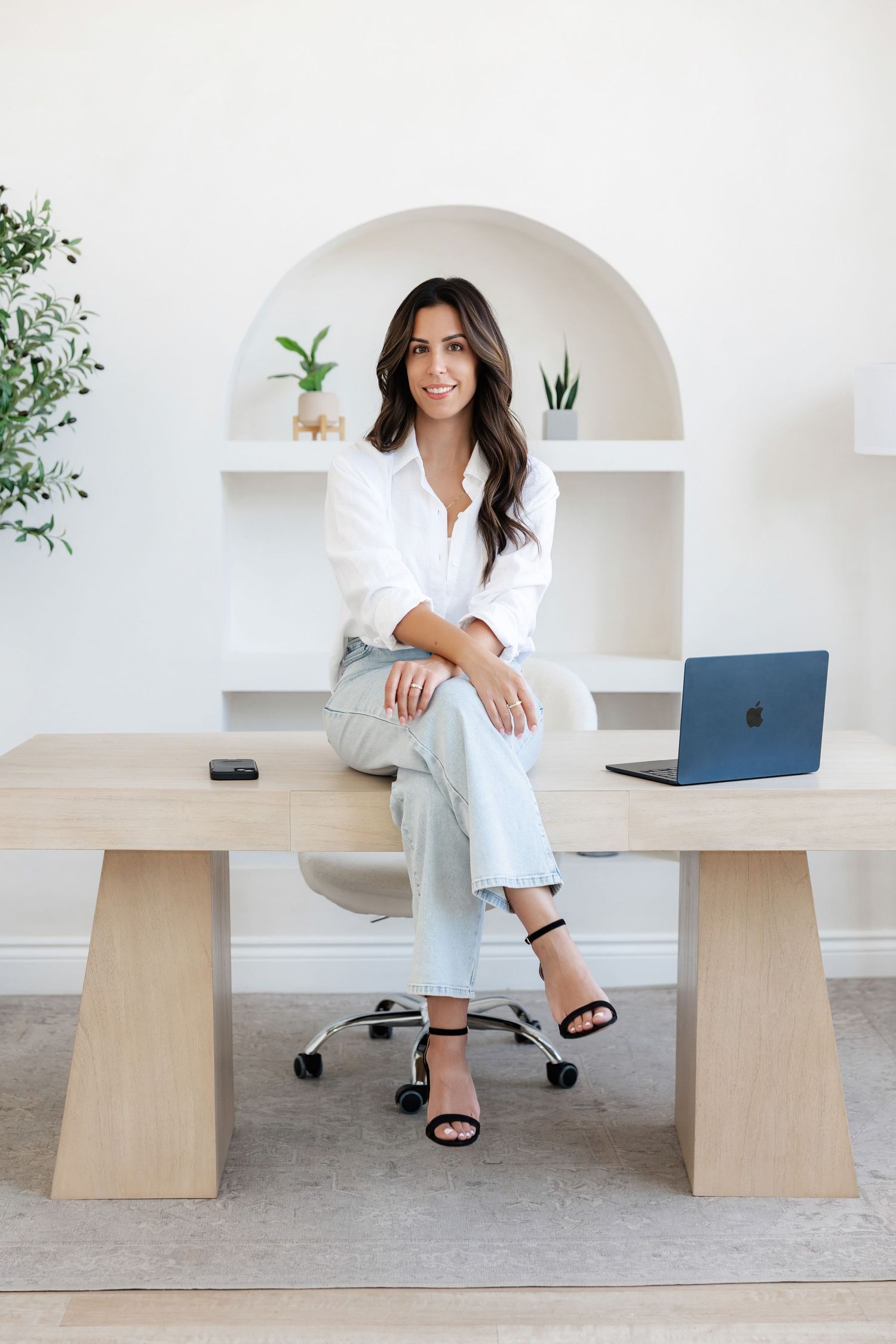 Woman in business casual attire sits at wooden desk in modern white office space.