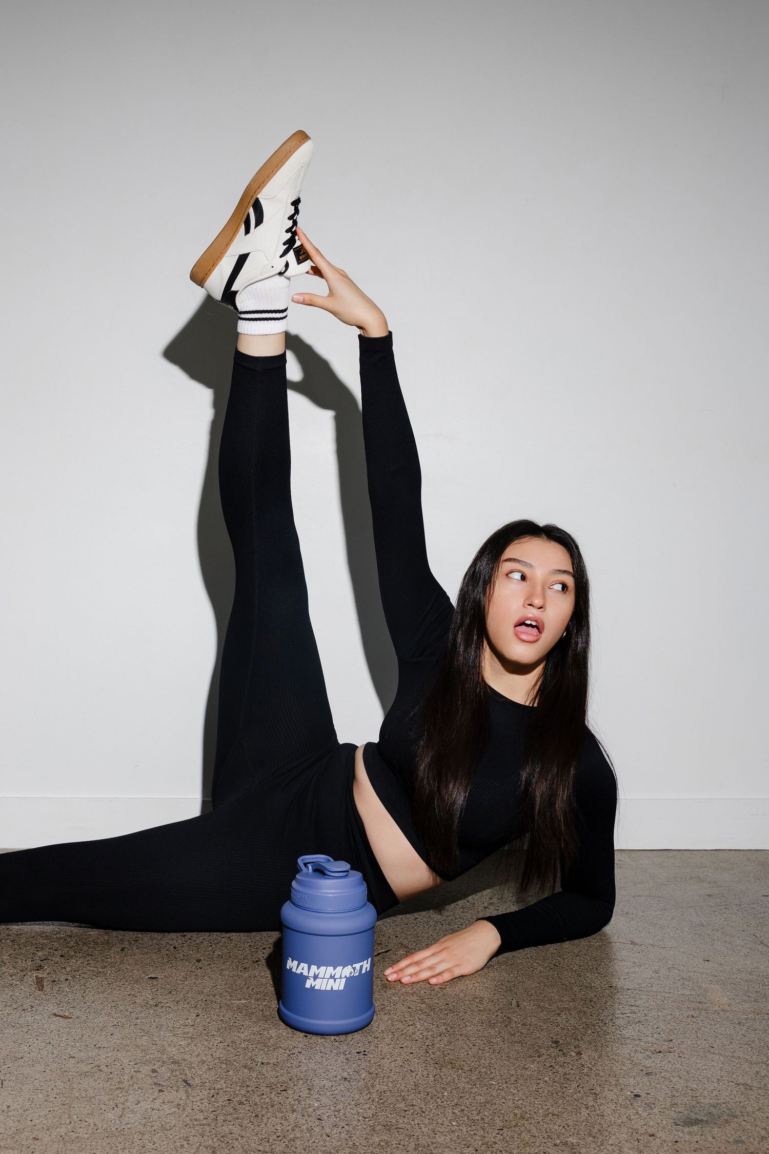 A person in black clothing lies on the ground posing playfully with a blue and white branded drink container.