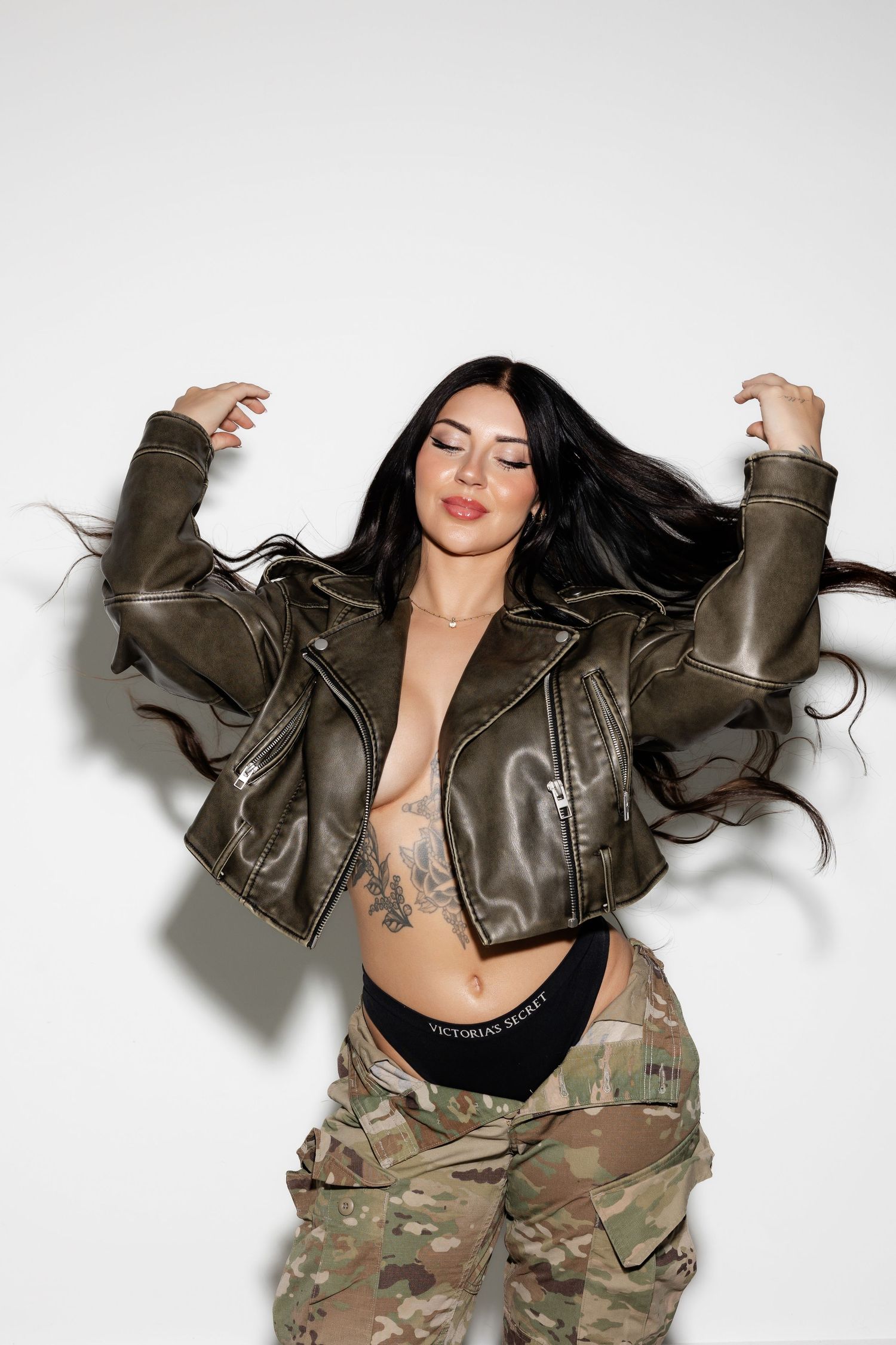 Model in leather jacket and camo pants poses energetically against white backdrop.