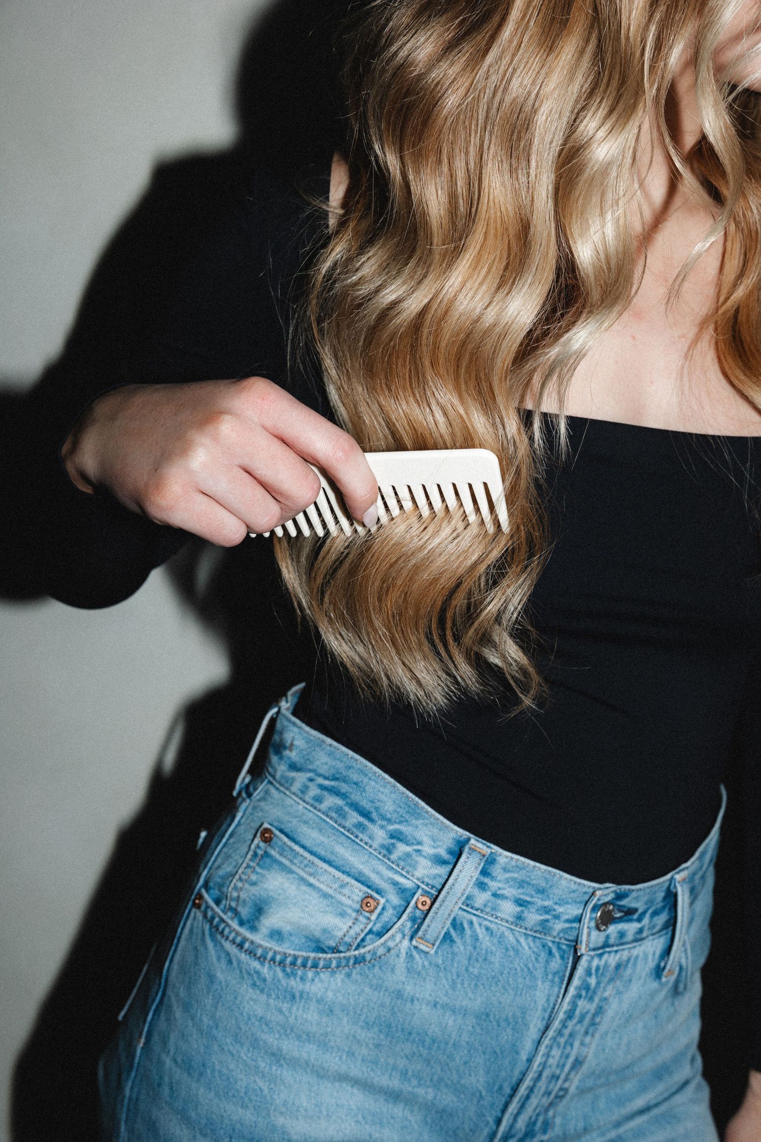 Styling wavy blonde hair with comb while wearing classic blue jeans.