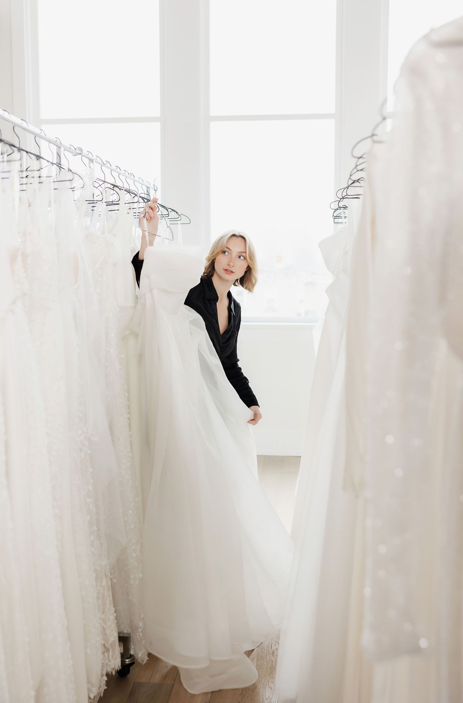 Staff members peeking through white wedding dresses hanging in a bright showroom.