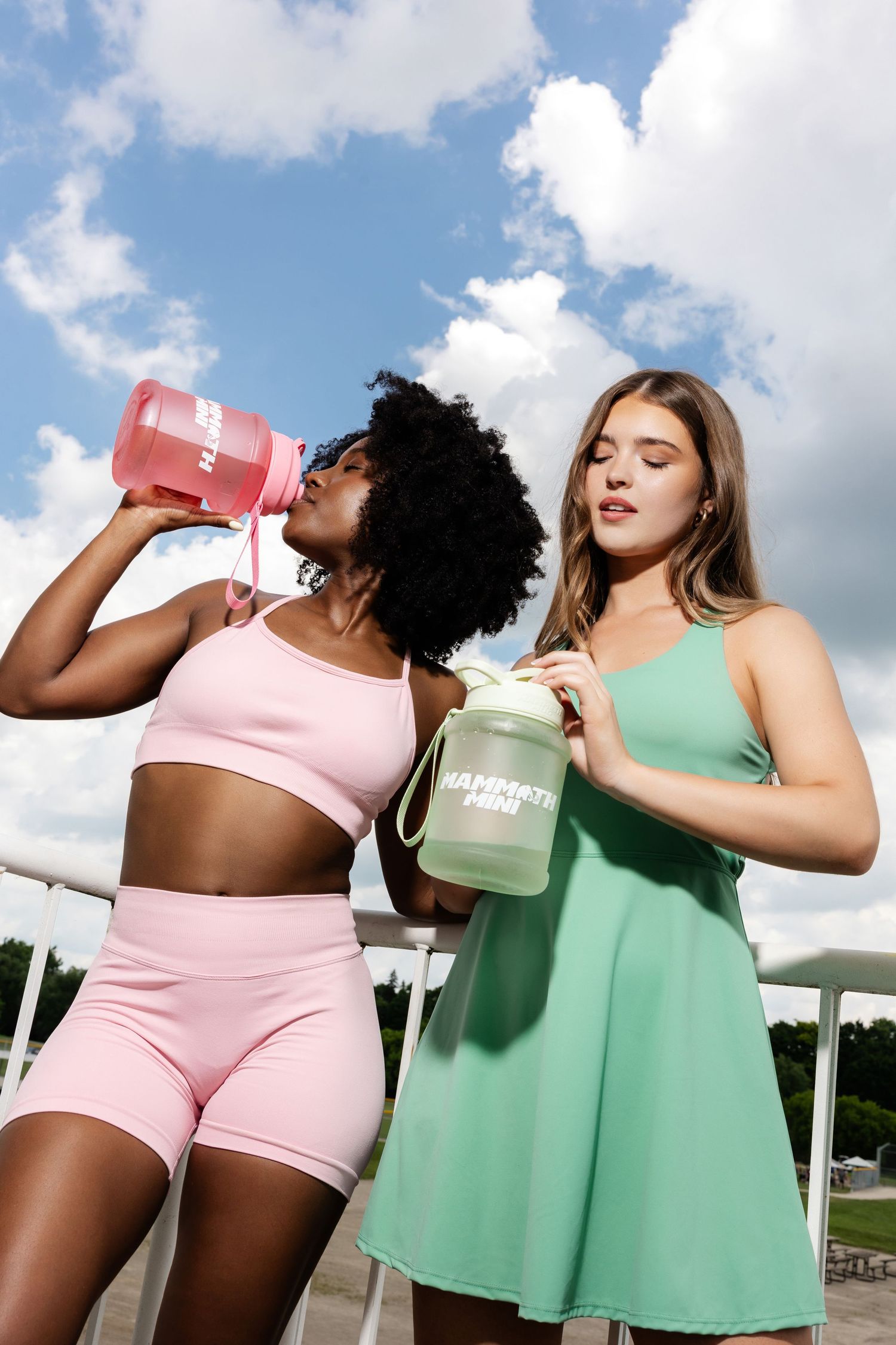Athletic models in pink and green outfits drinking from matching colored water bottles outdoors.