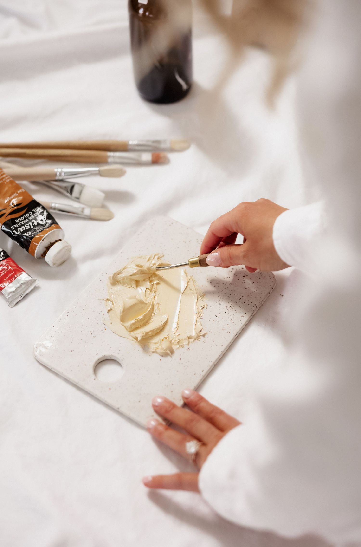 Close-up of hands mixing beige paint colors on white palette with brushes nearby.