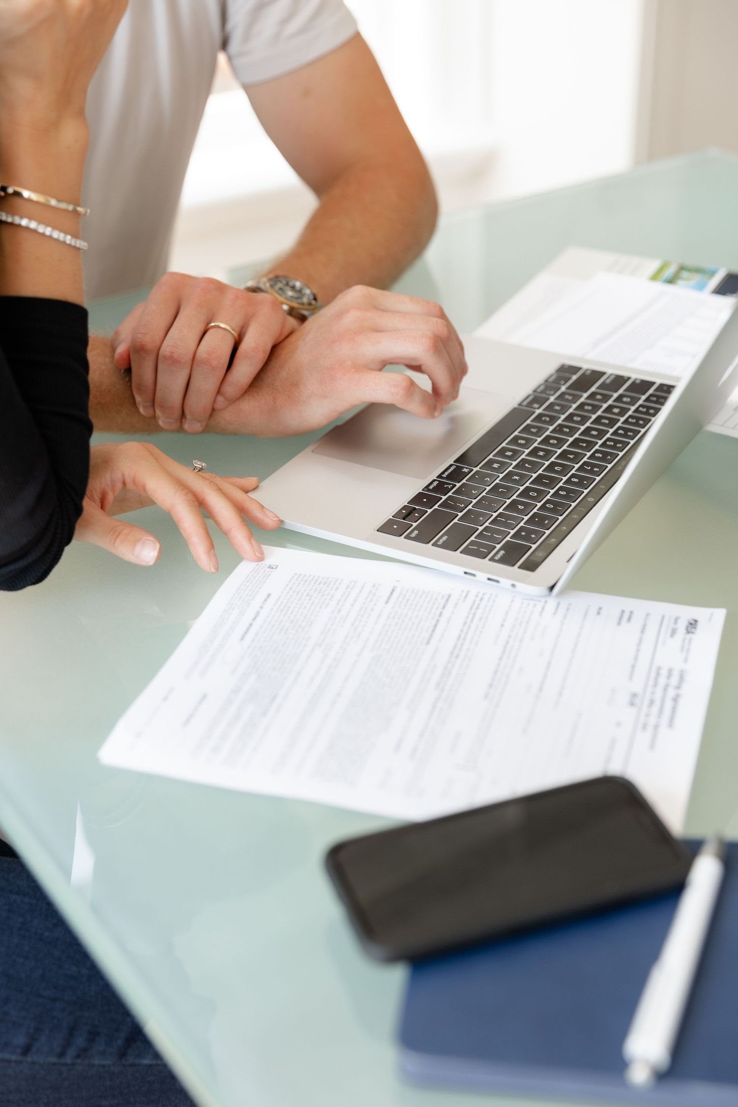 Hands reviewing documents and using a laptop on a glass desk surface.