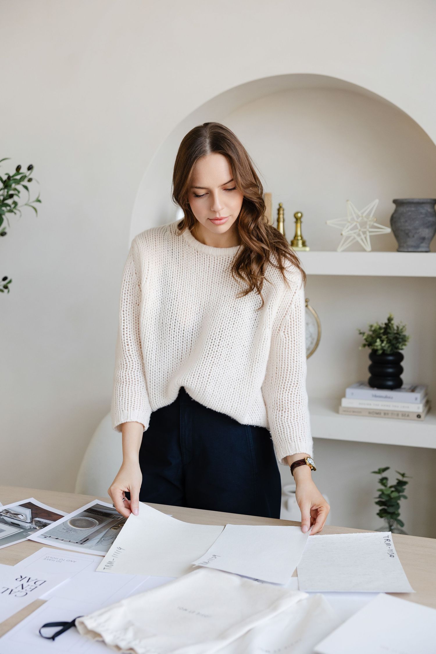 Designer in cream sweater reviews documents at desk with built-in shelving and decor in background.