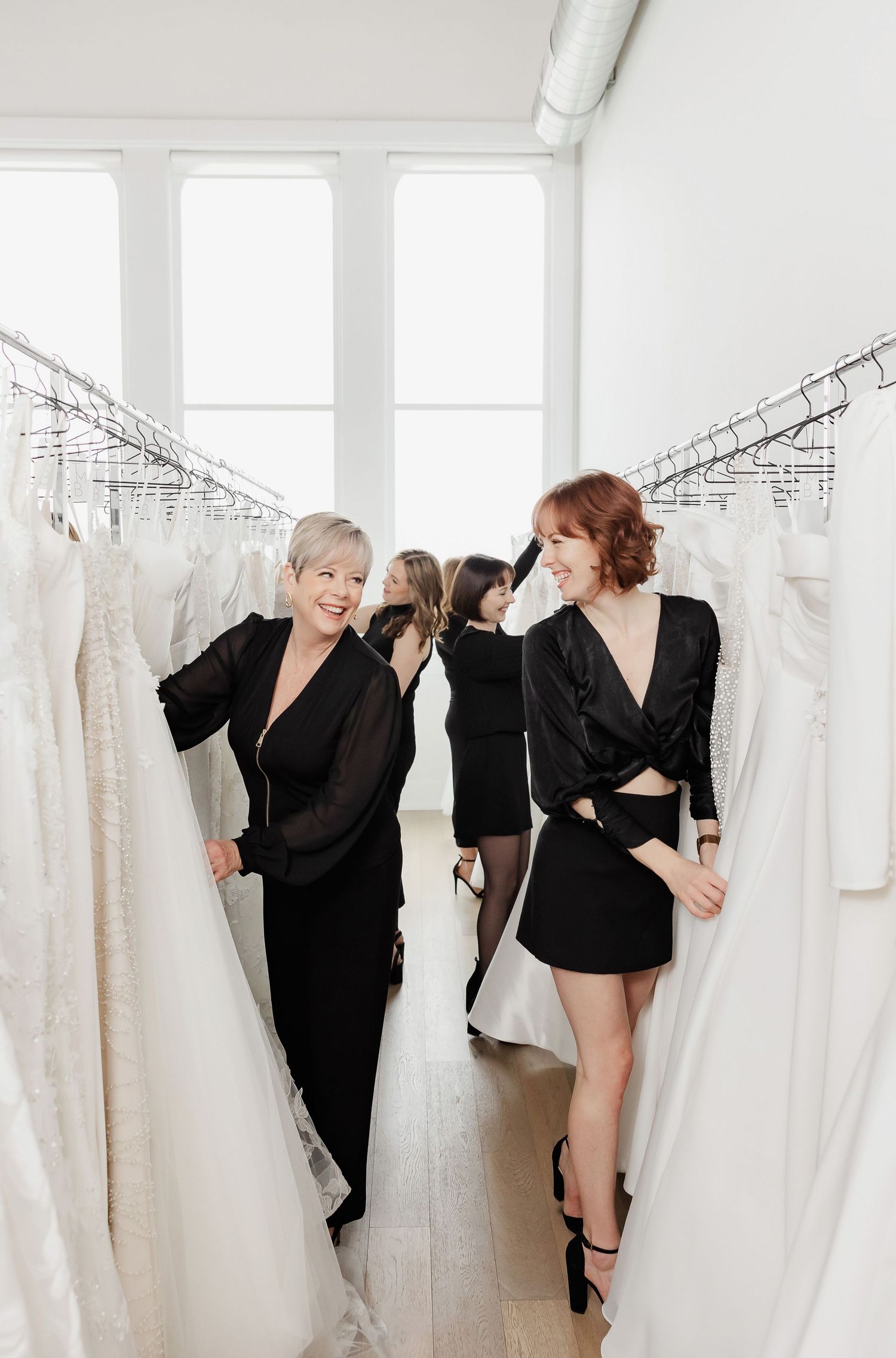 Team members playfully posing between racks of white wedding gowns in a bright showroom.