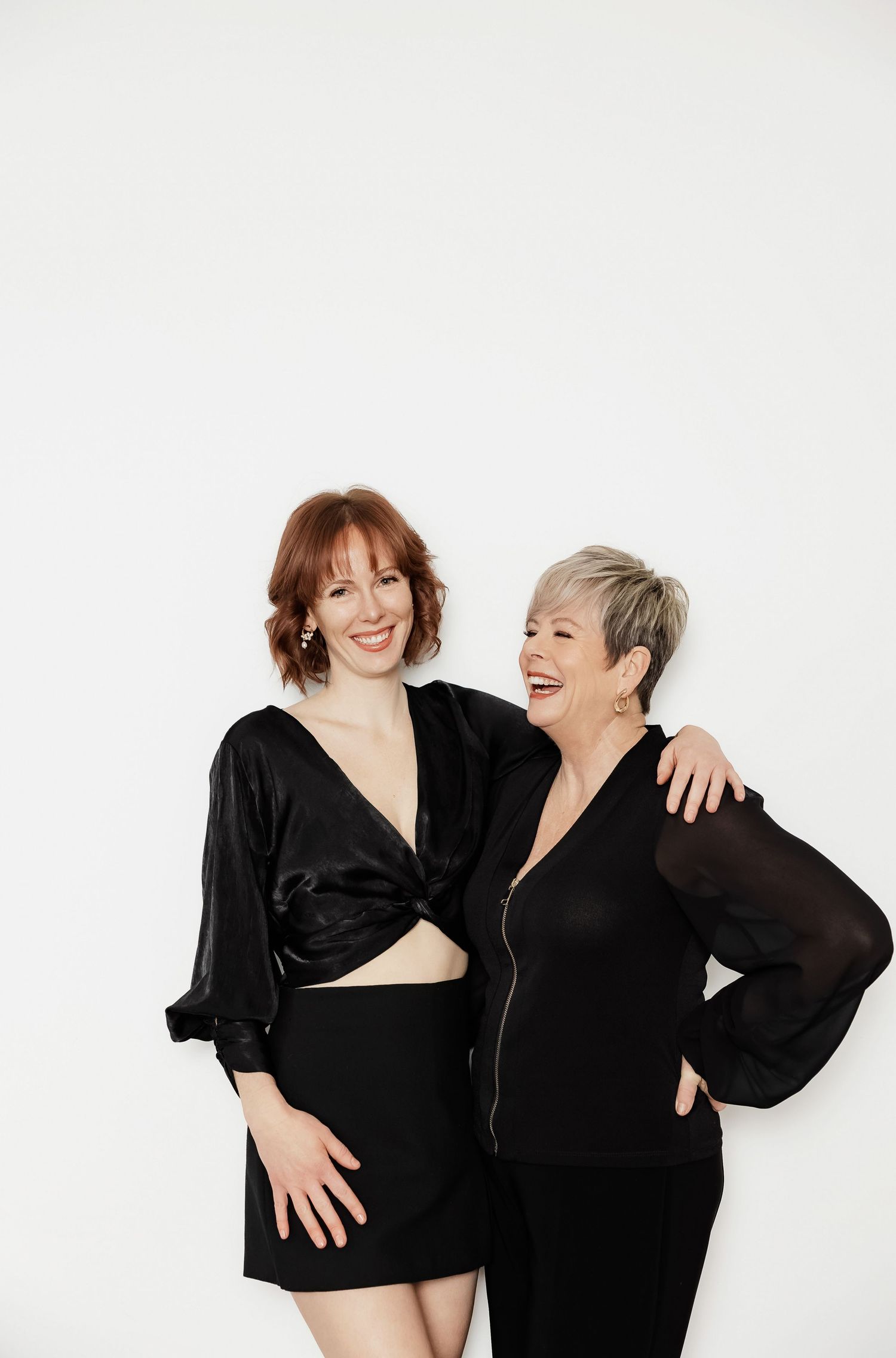 Two models in black outfits share candid laughing moment during photo shoot.