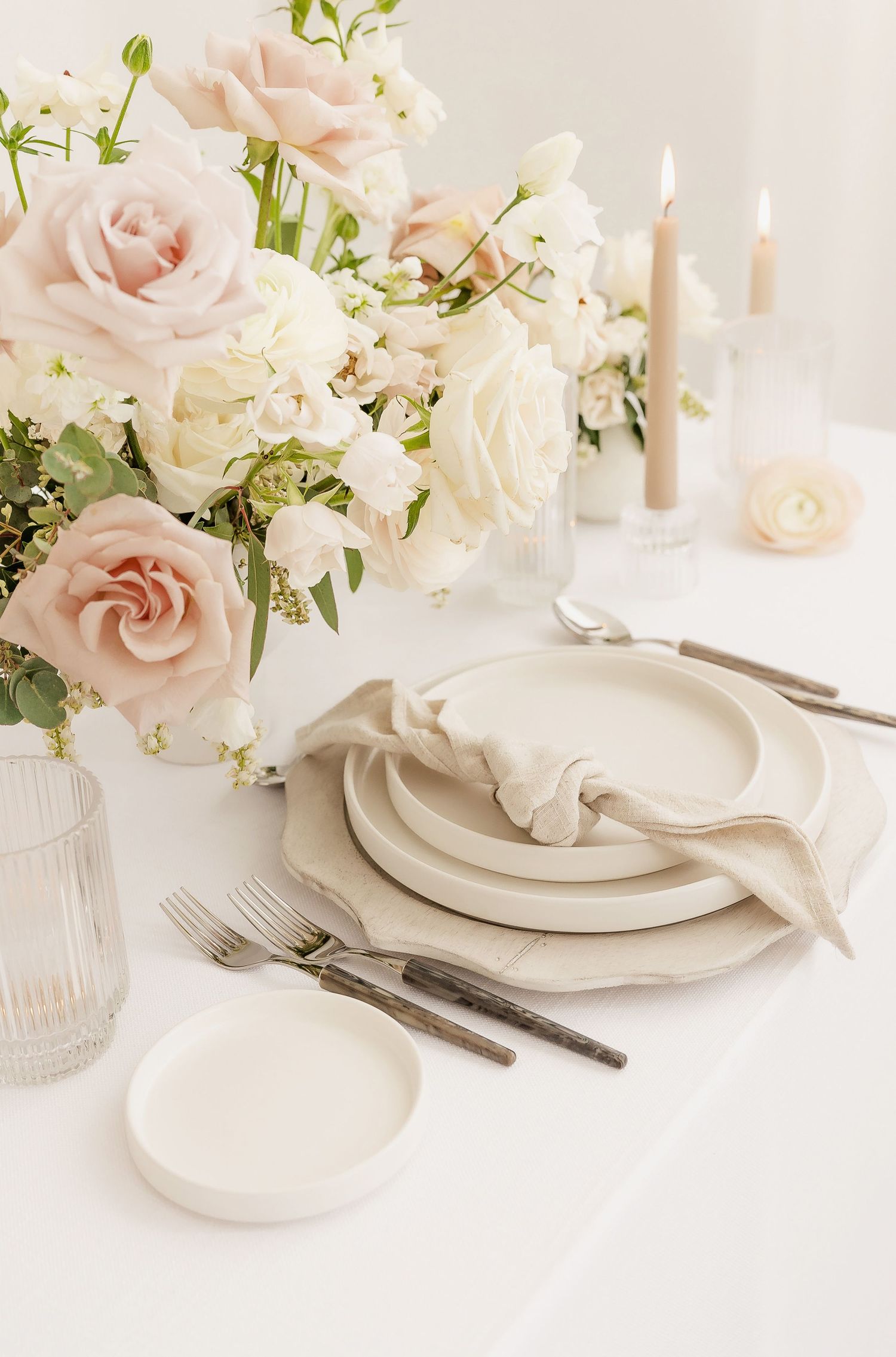 Elegant table setting with pink and white roses, candles, and vintage silverware on white linens.