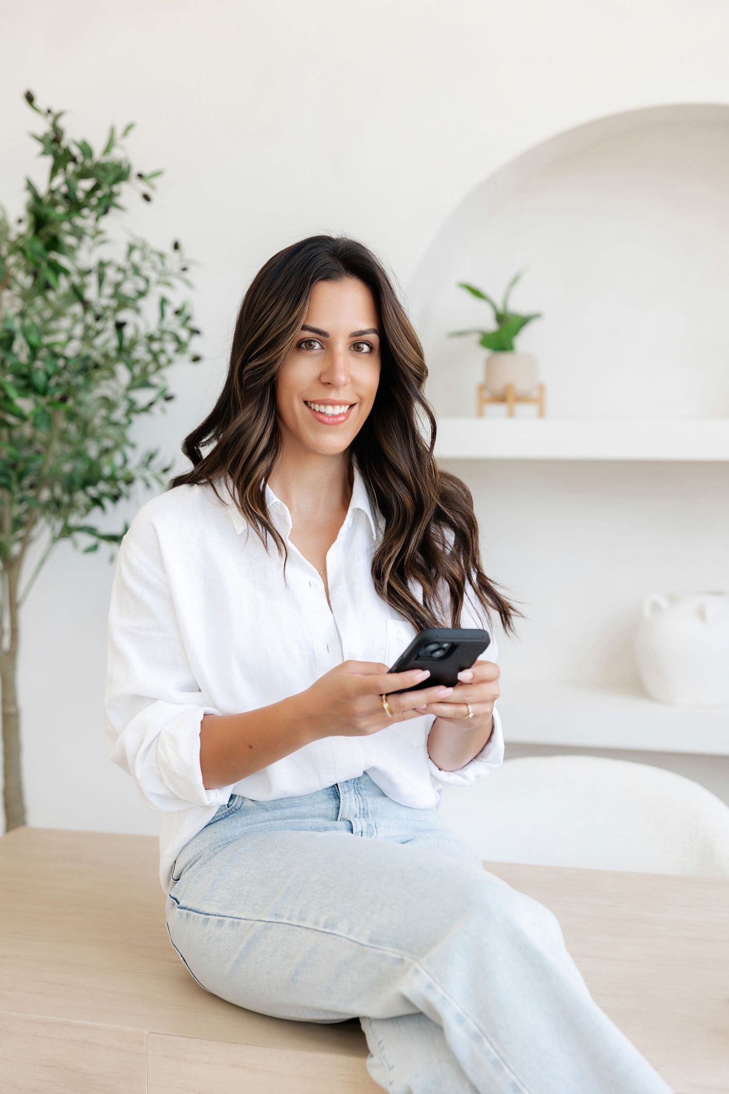 Woman in white button-down shirt holds phone while sitting at desk with greenery in background.