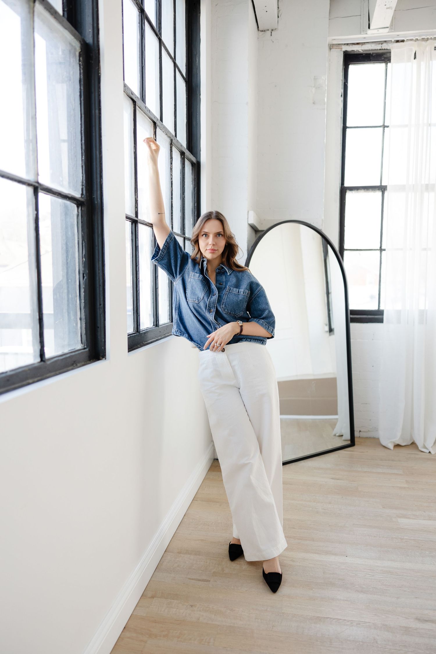 Dynamic pose against industrial windows wearing denim shirt and white trousers.