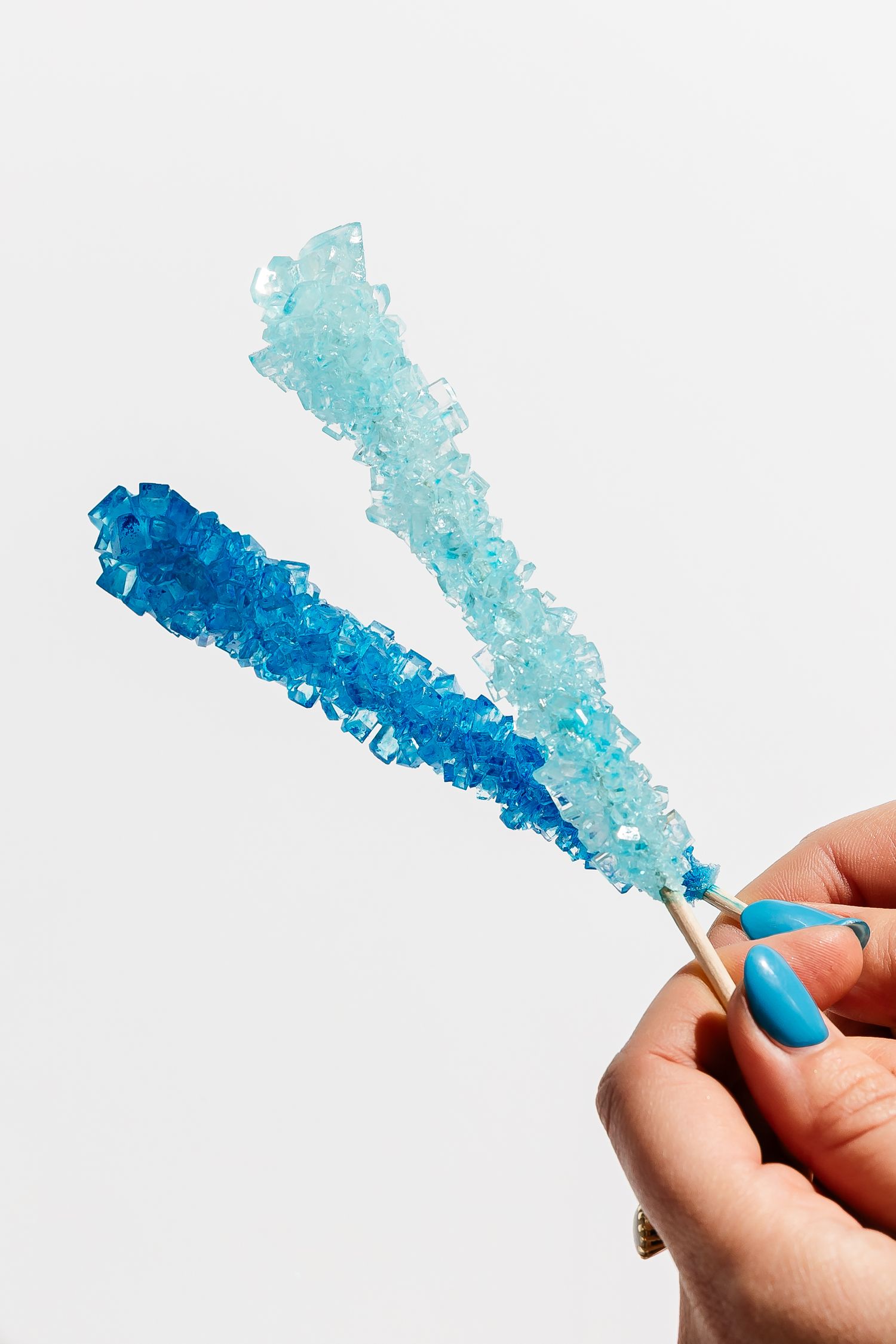 Hand holding blue rock candy crystals on stick against white background.