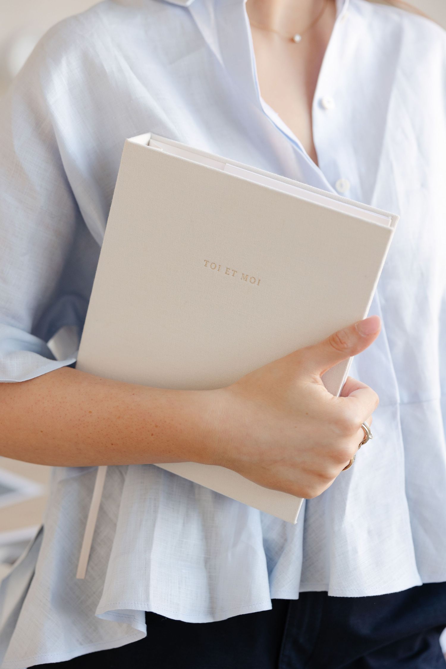 Person in white blouse holding cream-colored notebook against light background.