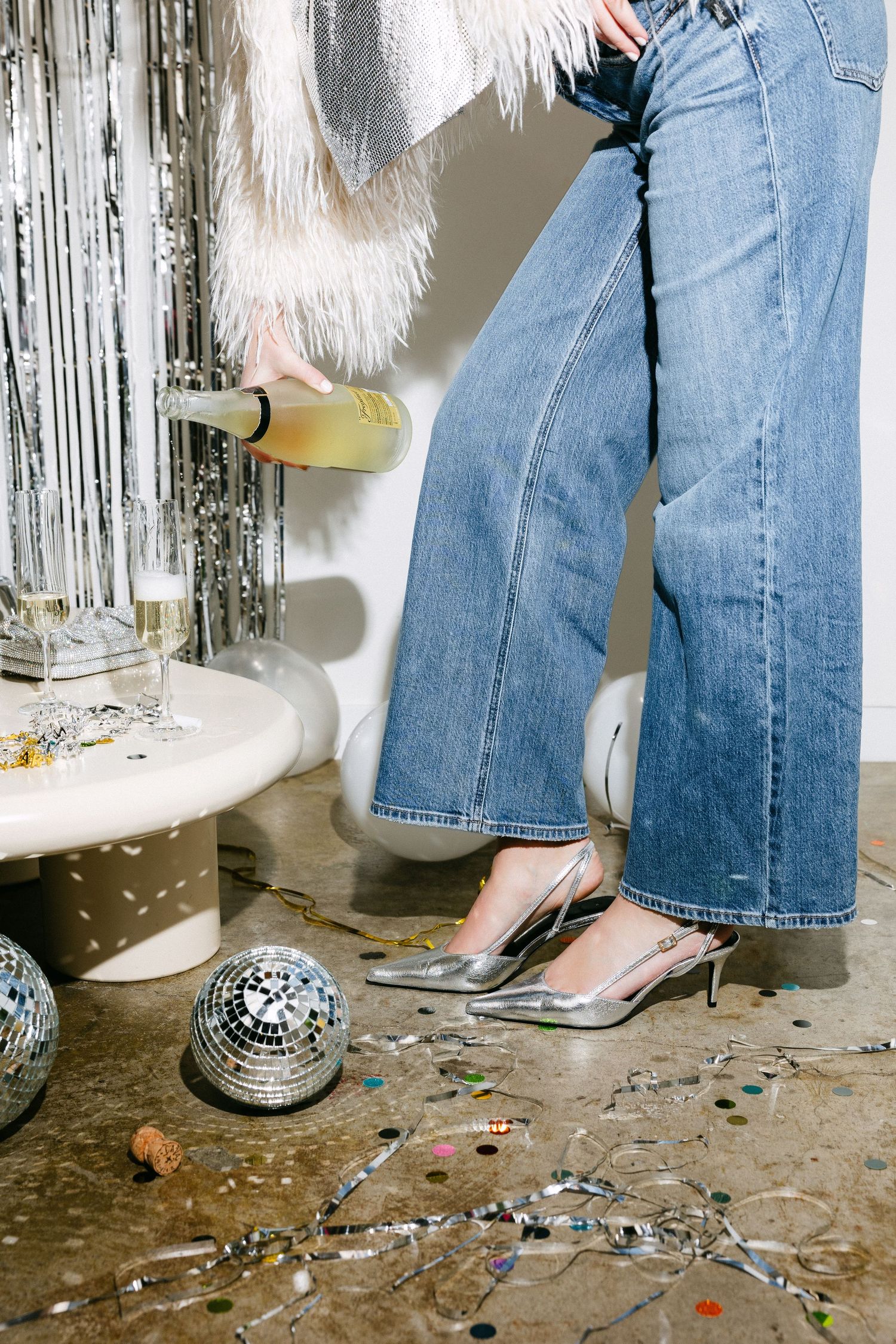 Close-up of silver kitten heels and jeans with disco ball and confetti on floor.