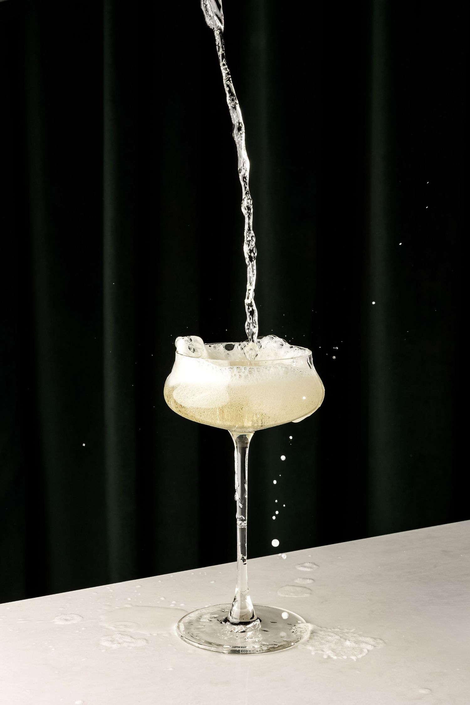 A stream of liquid being poured into a coupe glass creating an artistic splash against a dark backdrop.