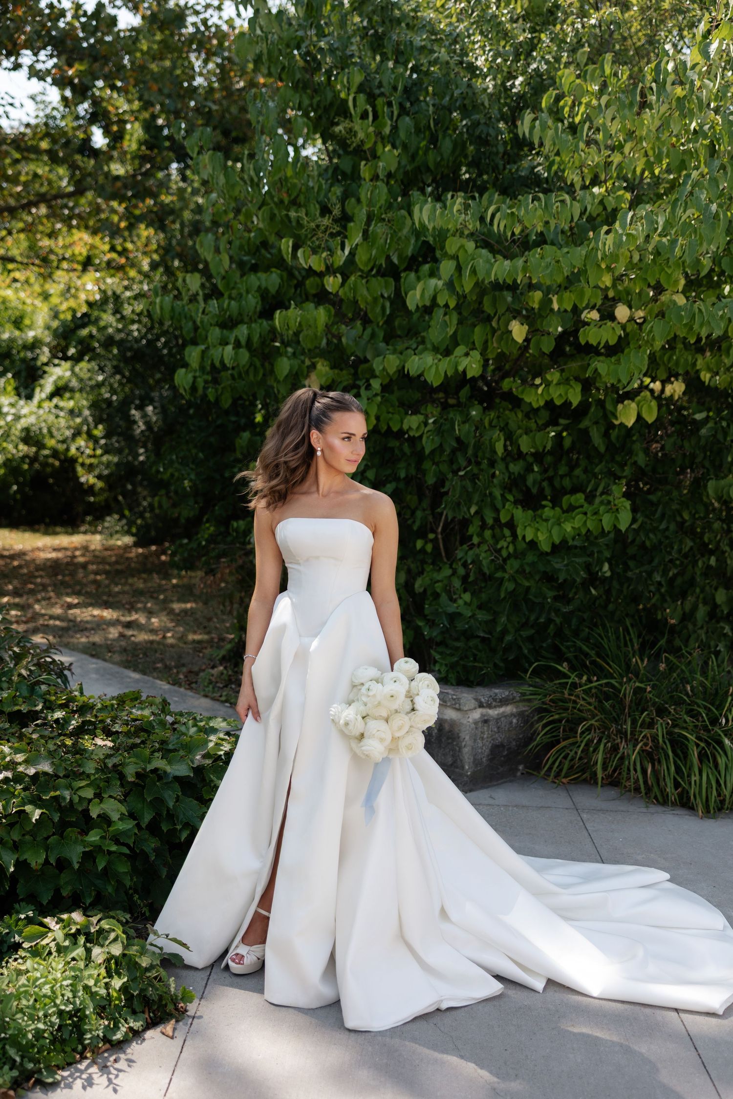Bride in elegant strapless wedding gown with thigh slit holding white bouquet in garden setting.