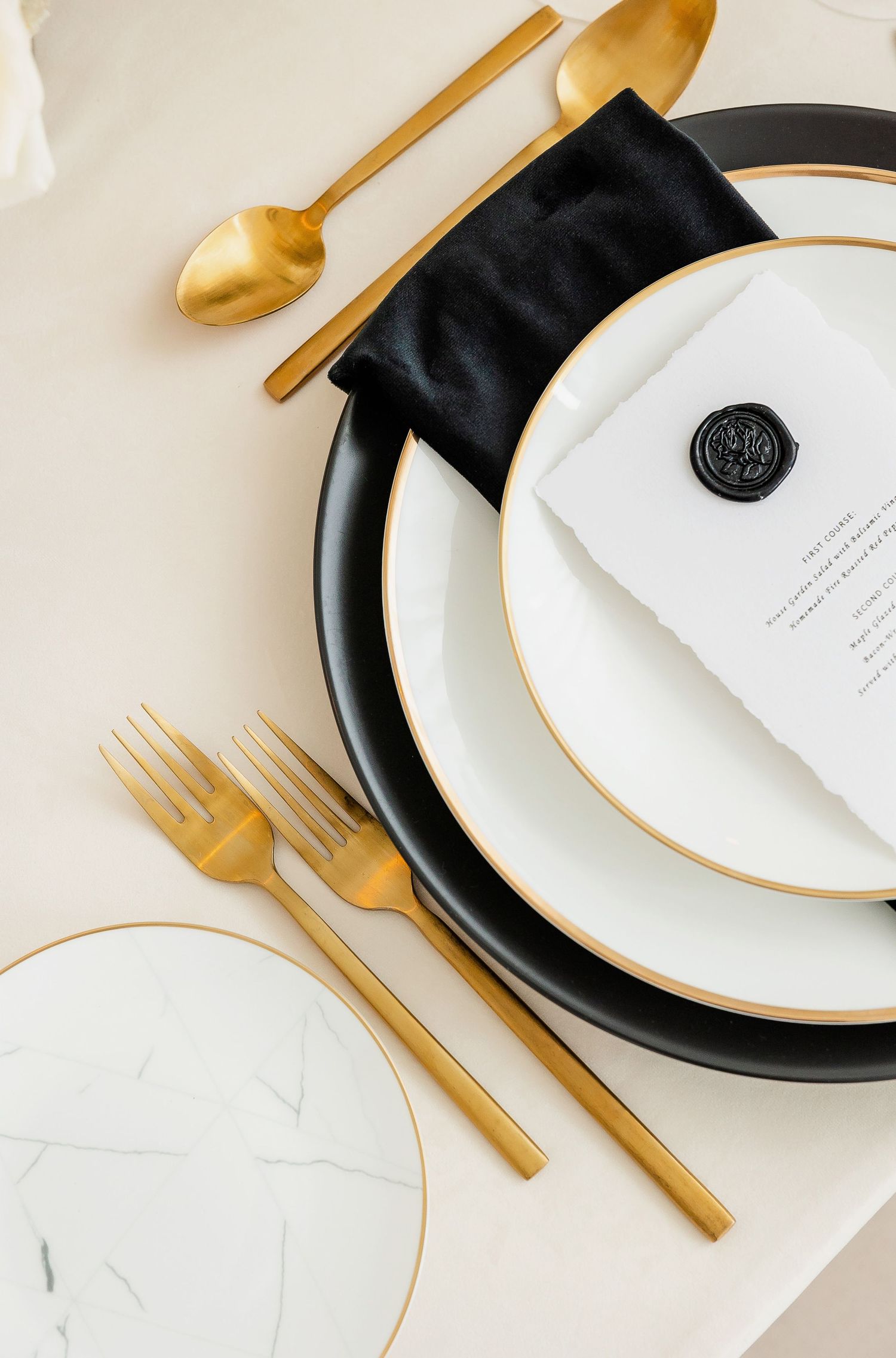 Minimalist table setting featuring black and white plates with gold accents and wax seal menu.