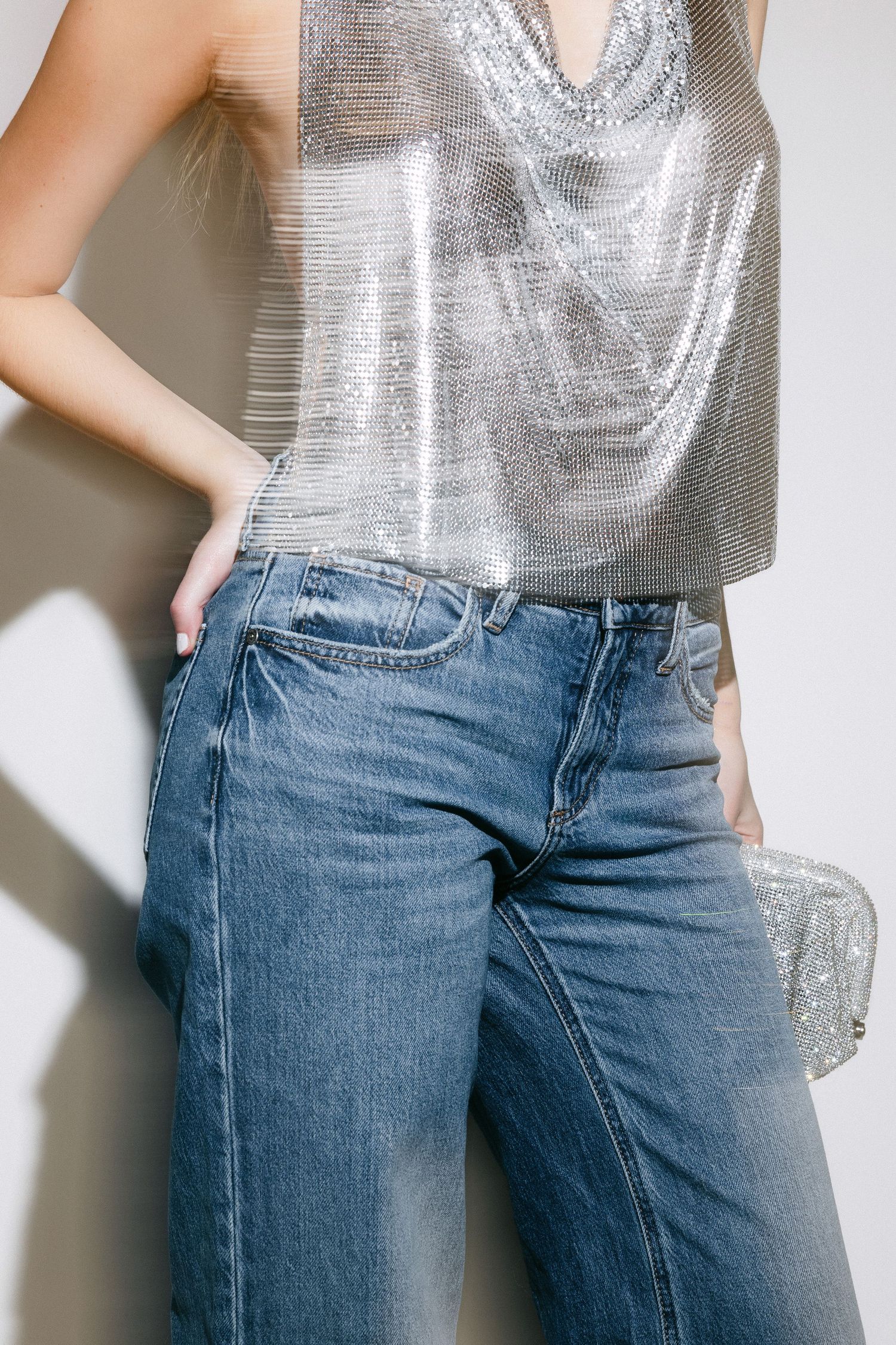 Woman wearing silver metallic top and blue jeans poses against white wall.