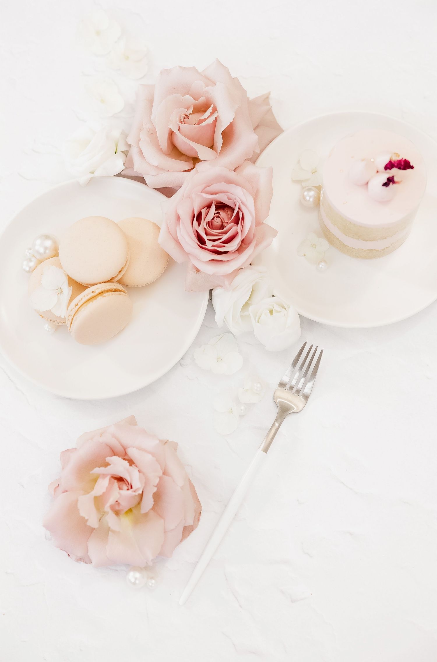 Elegant table setting with pale pink roses, macarons and desserts on white plates with delicate details.
