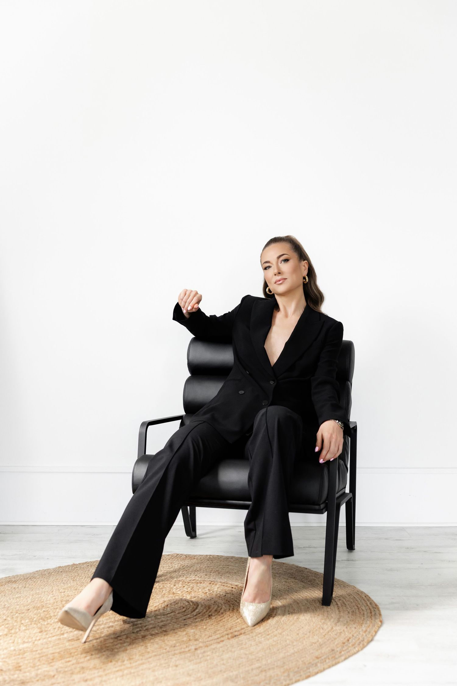 Woman in black pantsuit reclines elegantly in modern black chair on round beige rug.