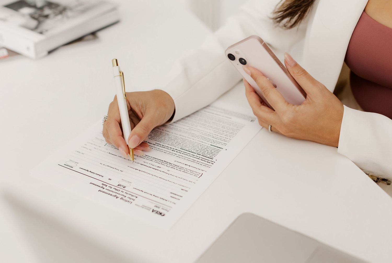 Close up of hands signing document while holding phone at white desk.