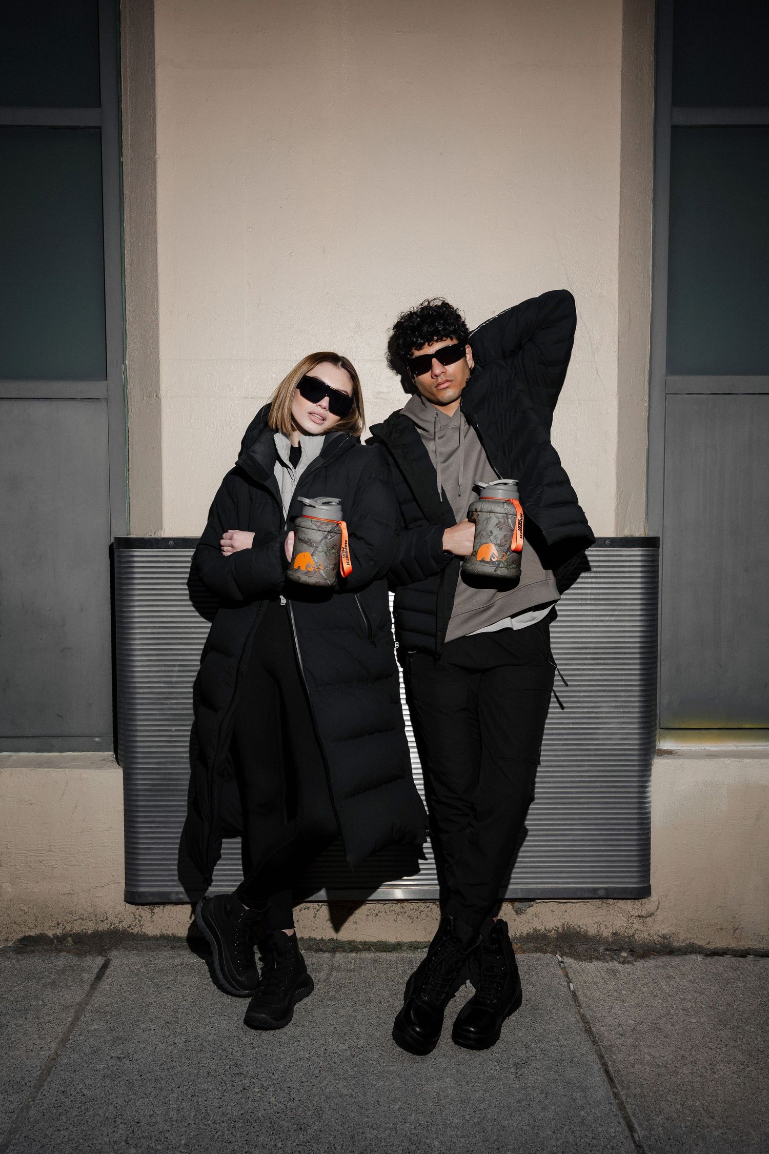 Two people dressed in black sit casually against a light colored wall outdoors.