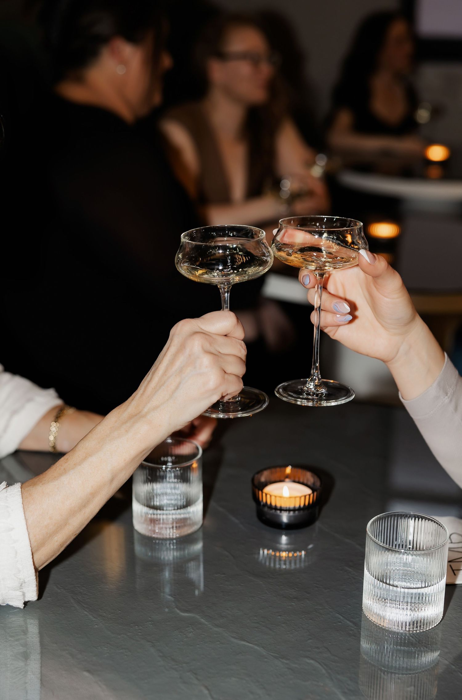 Hands reaching across a dark table to toast with champagne glasses by candlelight.