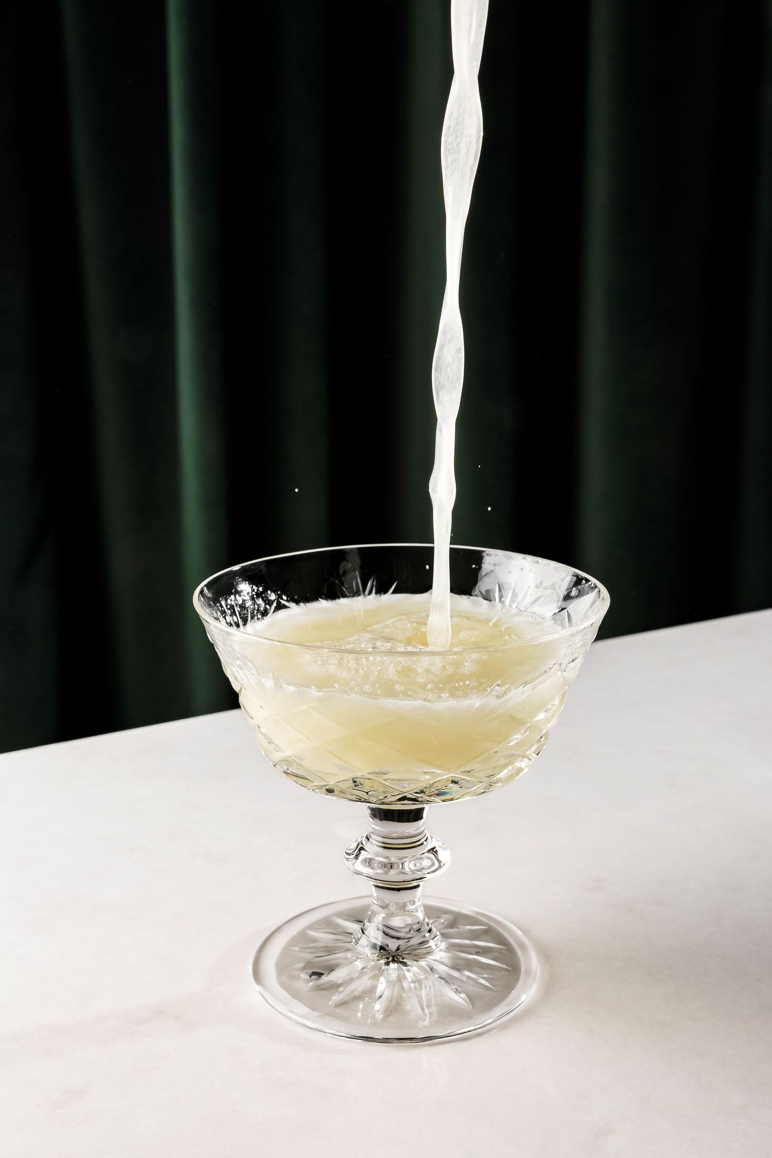 A white liquid stream pouring into a vintage coupe glass on a white surface.