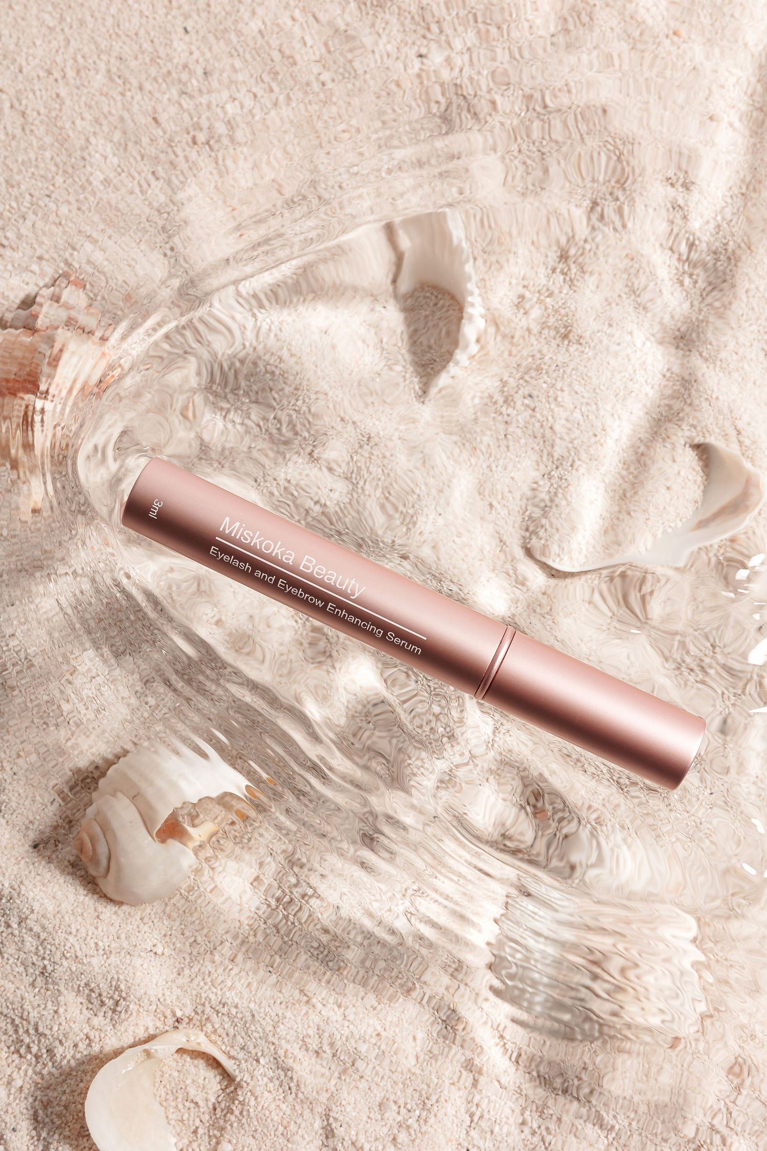 Rose gold beauty product tube lying on sandy textured surface with water splashes.