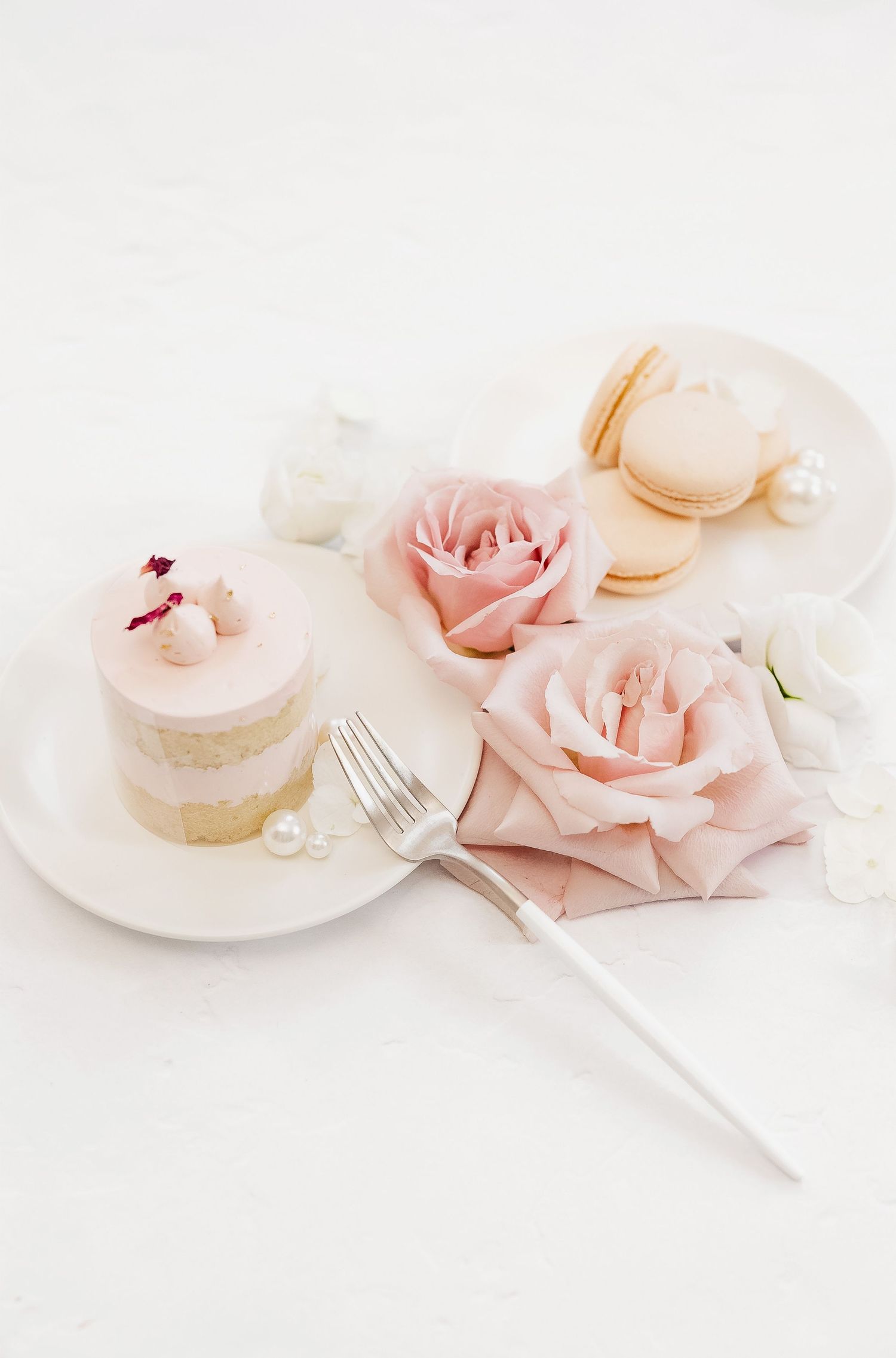 Delicate pink roses and macarons with a small cake decorated with rose petals on white plates.