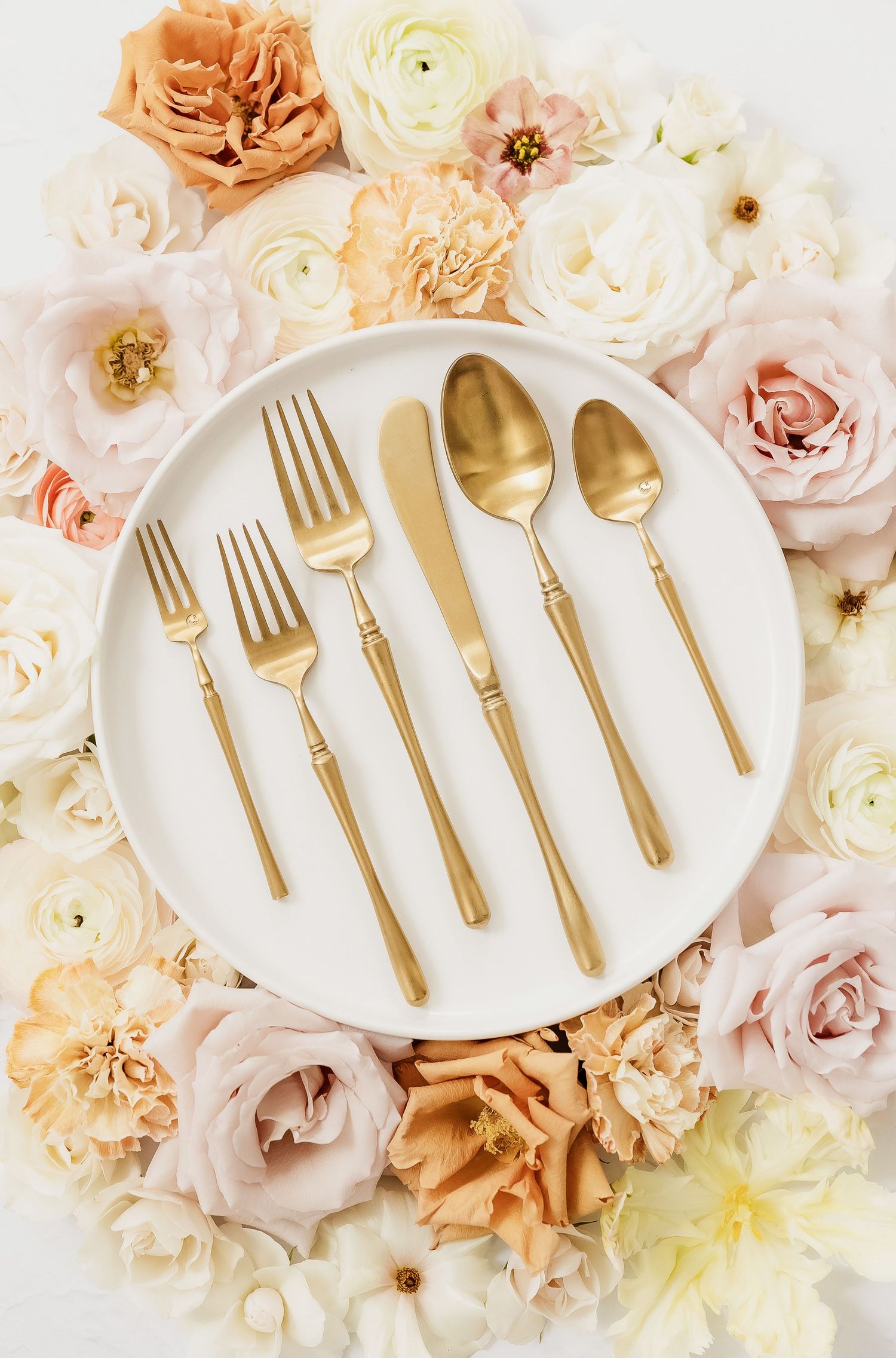 Gold flatware displayed on white plates surrounded by peachy pink and cream roses.