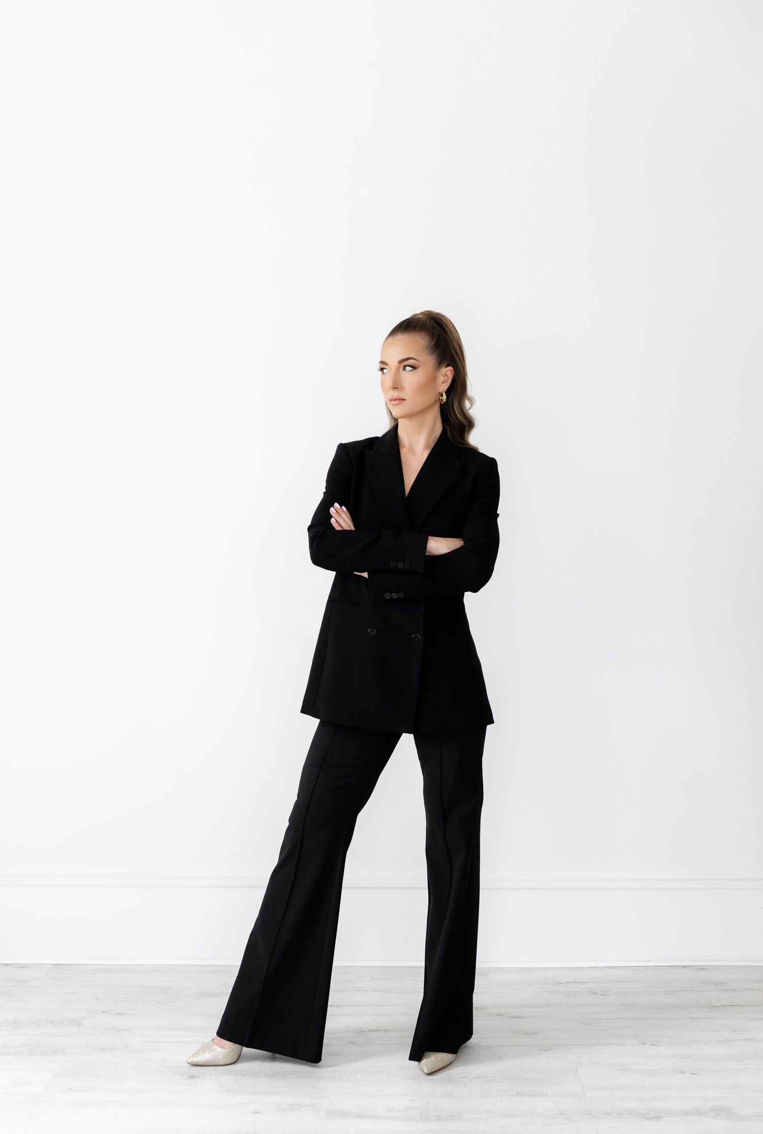 Woman in black business suit stands confidently with arms crossed against white background.