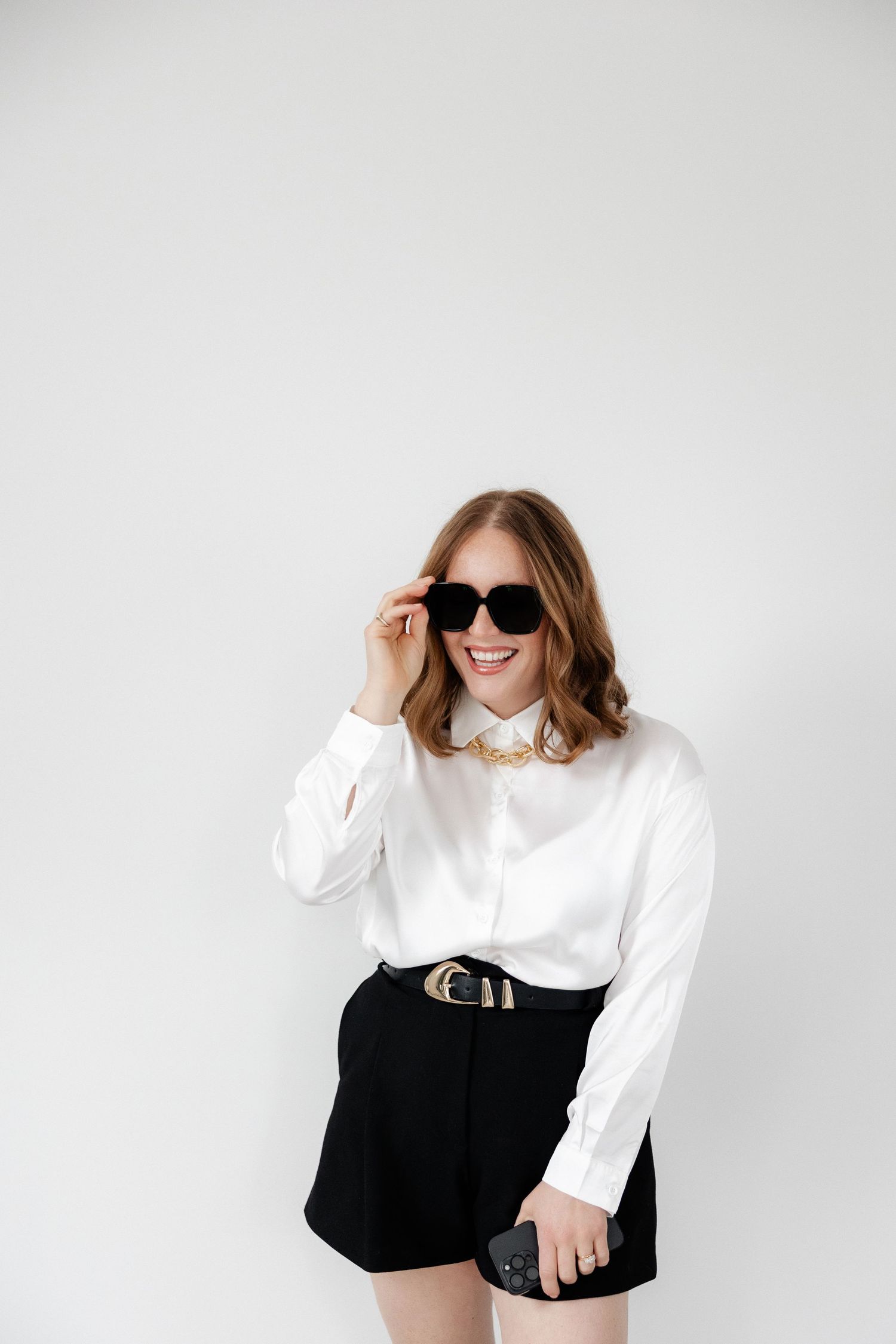 Stylish outfit featuring white blouse and black shorts with sunglasses against white background.