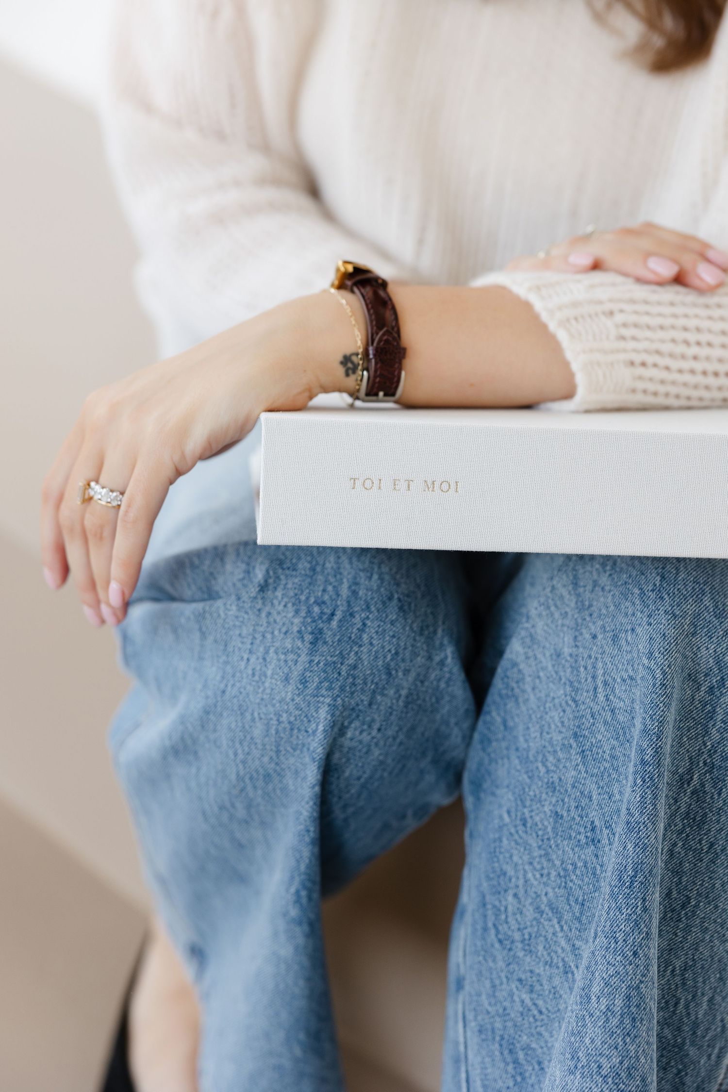 Person wearing blue jeans sits with white book or notebook on their lap in soft natural lighting.