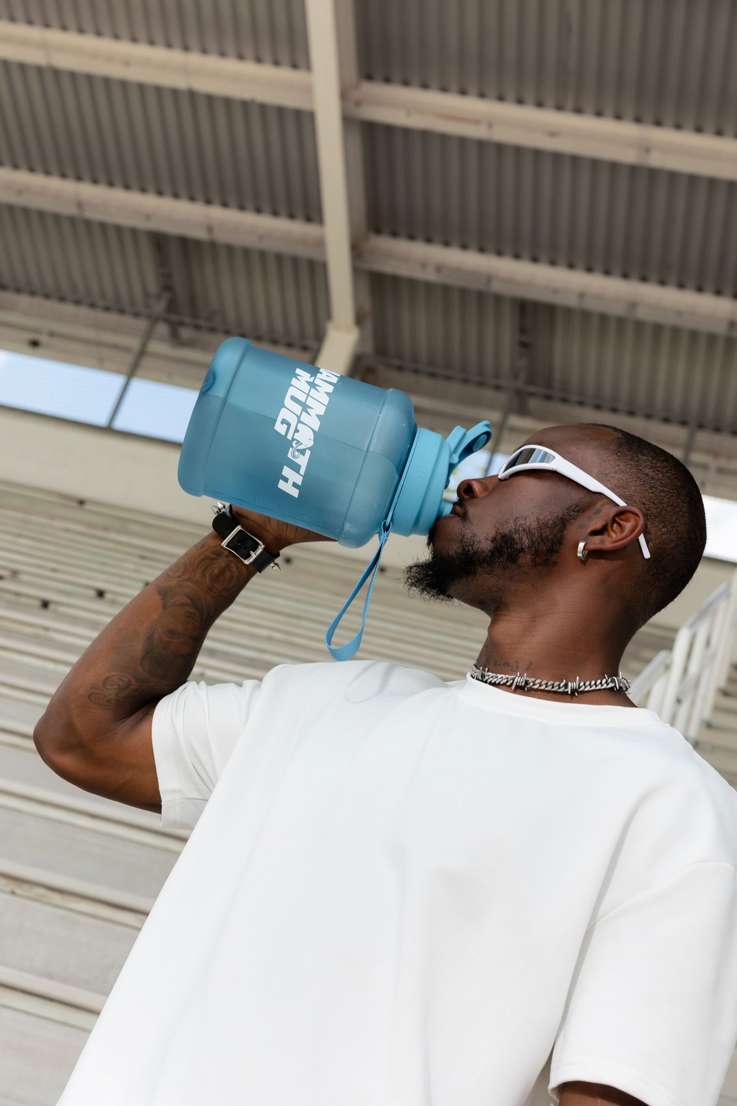 Person in white clothing drinking from large blue water bottle while seated on concrete bleachers.